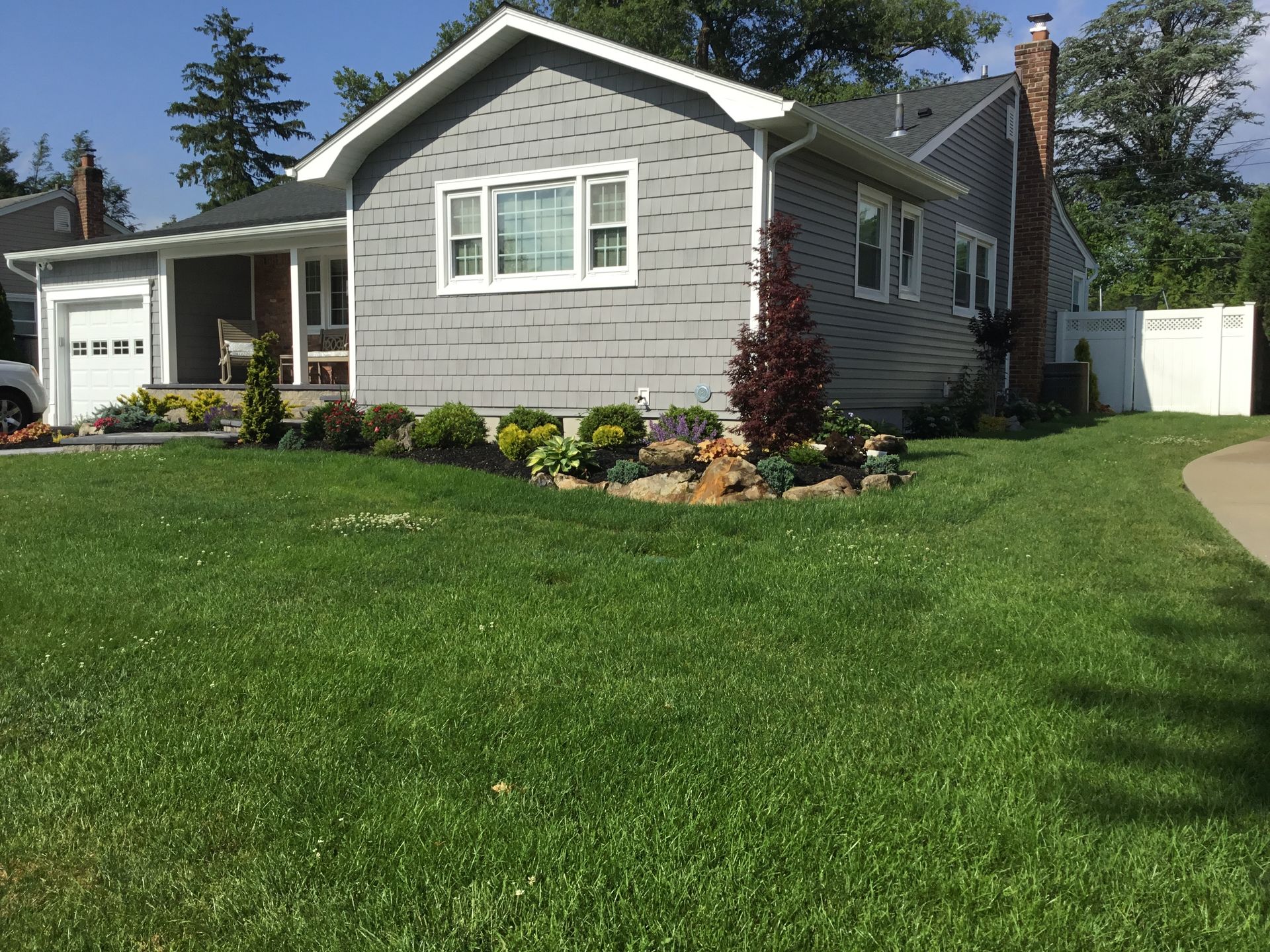 A single-story gray house with a front porch, large green lawn, and landscaped flower beds on a sunny day.