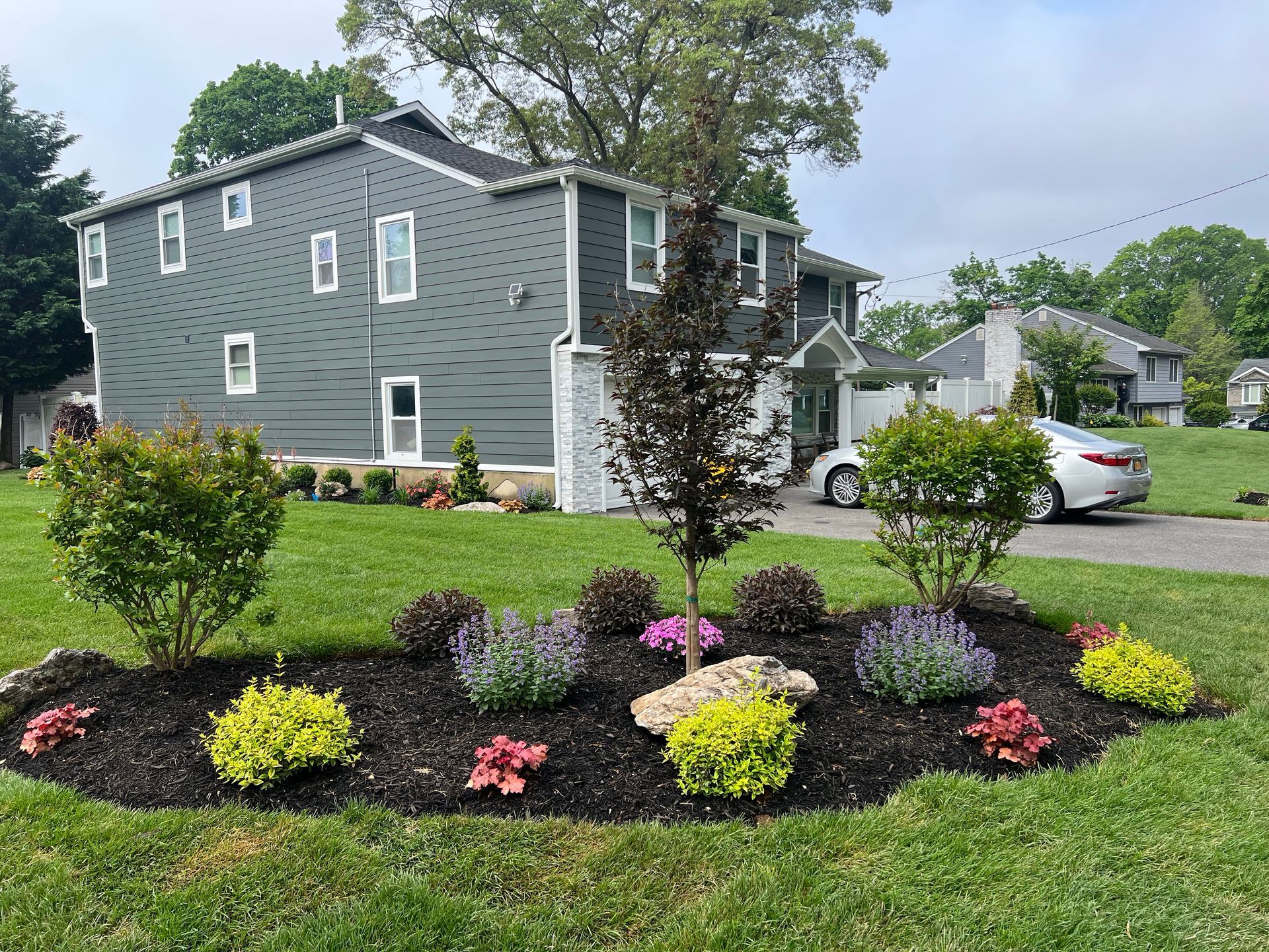A landscaped garden bed with mulch, shrubs, flowers, and a small tree in front of a gray two-story house.