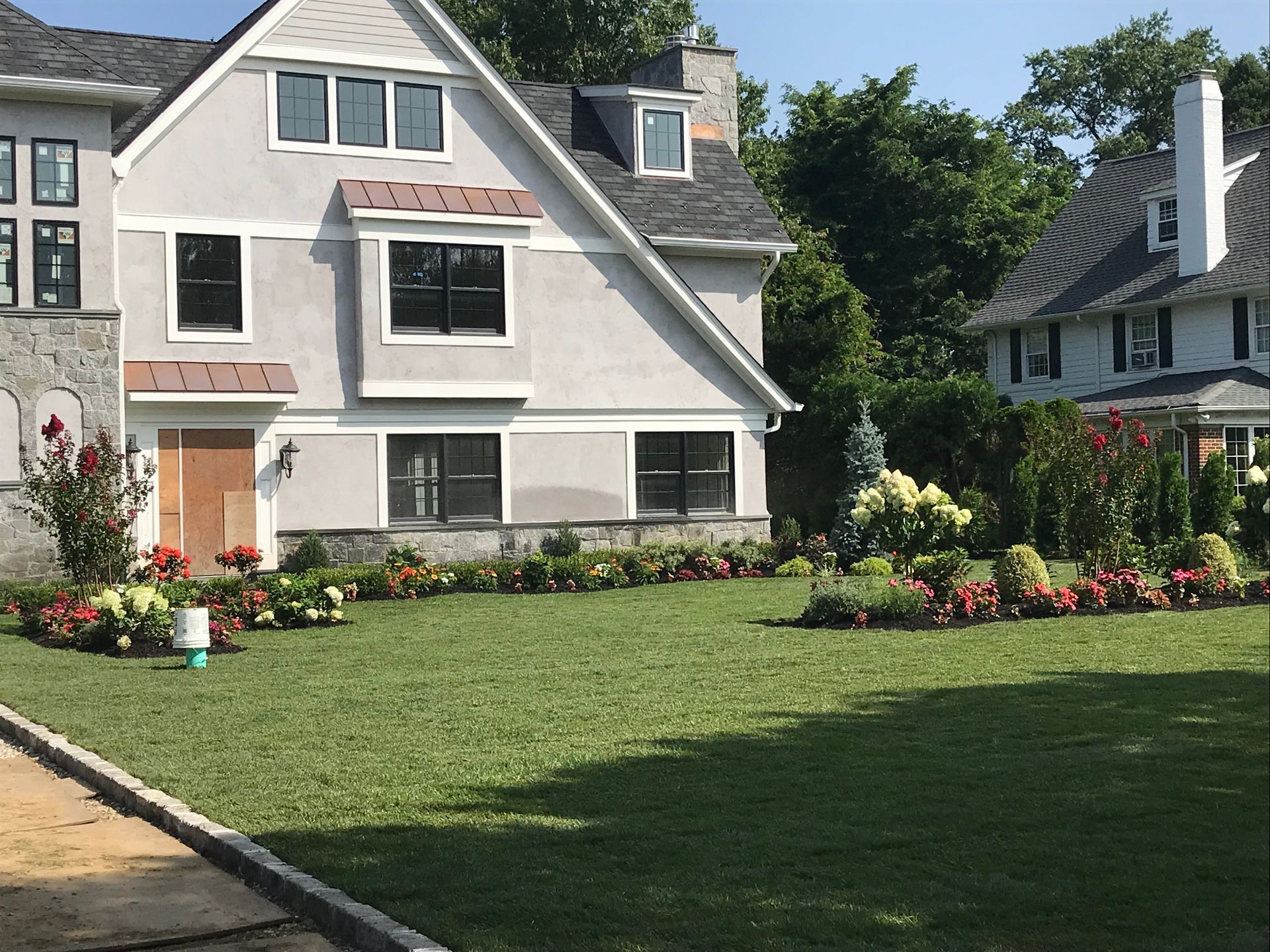 A two-story light gray house with a stone base and black-trimmed windows, fronted by a large lawn with colorful flowerbeds.