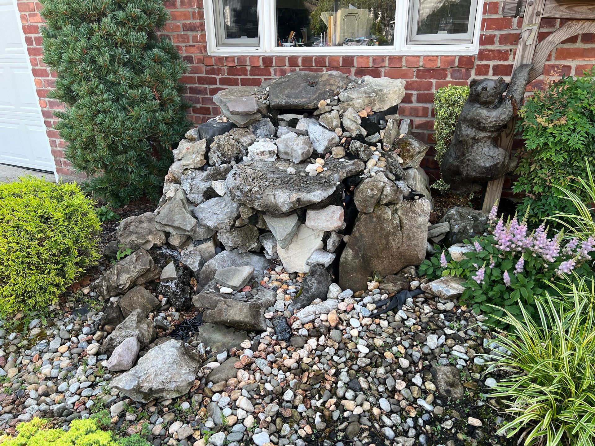 A rock garden feature with a small waterfall near a brick house wall, featuring a decorative bear statue and greenery.