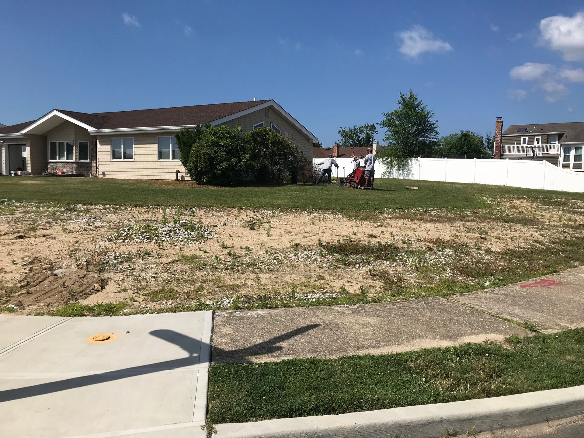 A sunny day view of a vacant, sandy residential lot next to a house with a white fence, with two people working nearby.