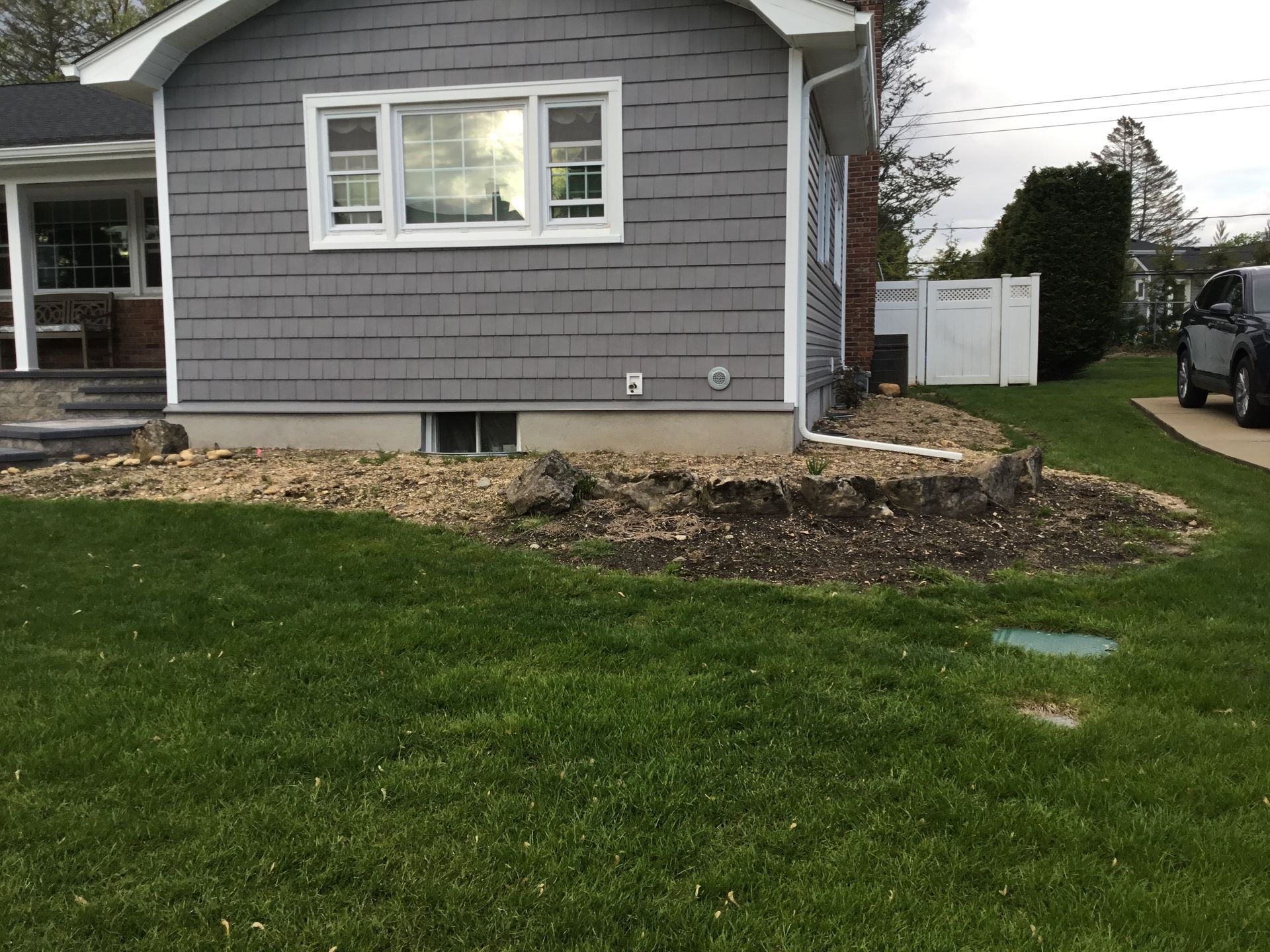 A gray shingled house with a window, basement vent, and a rock-covered garden bed in the front yard.