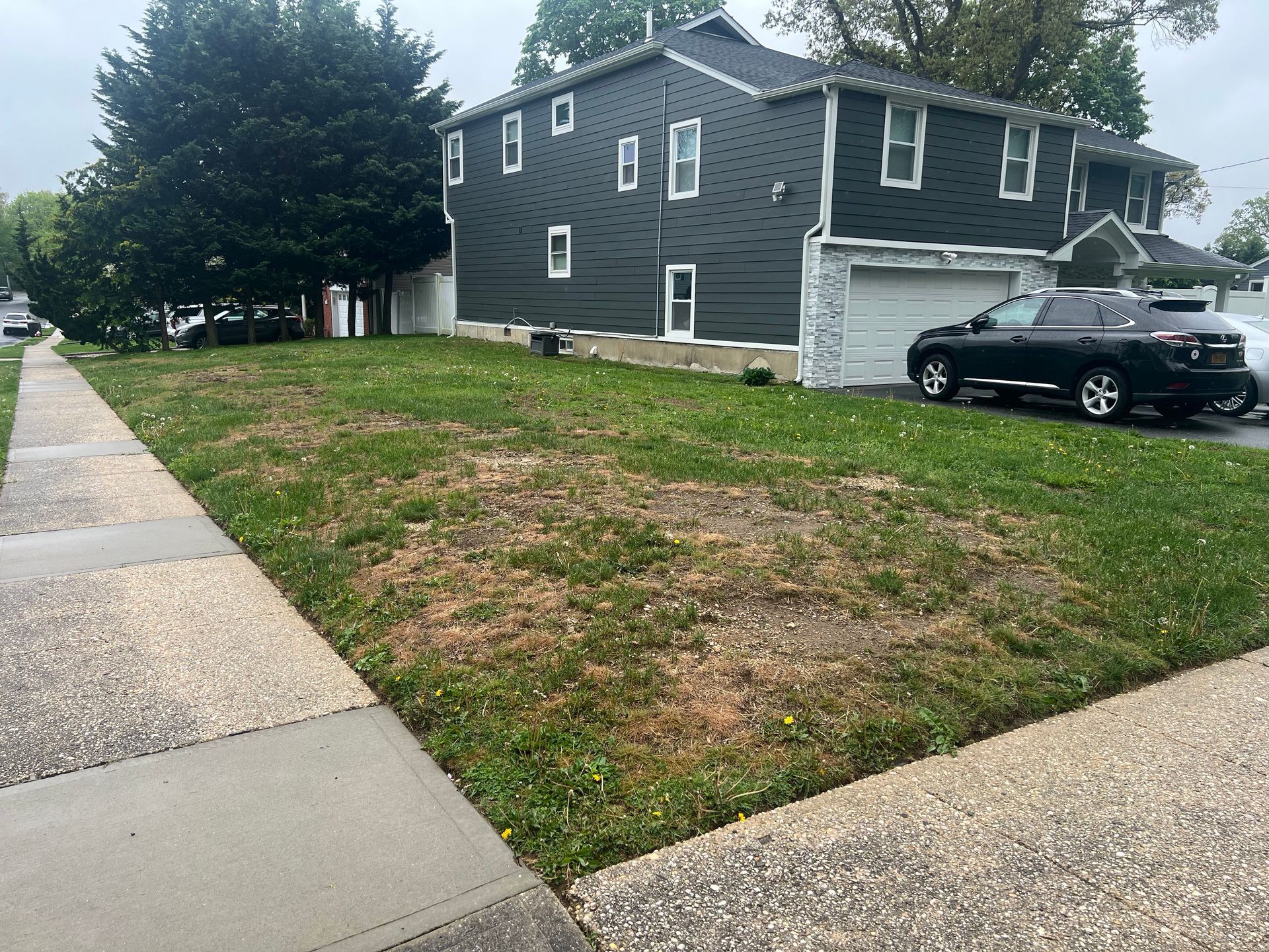 A gray two-story house with a driveway and car, seen from a sidewalk next to a patch of grass with brown, dry areas.
