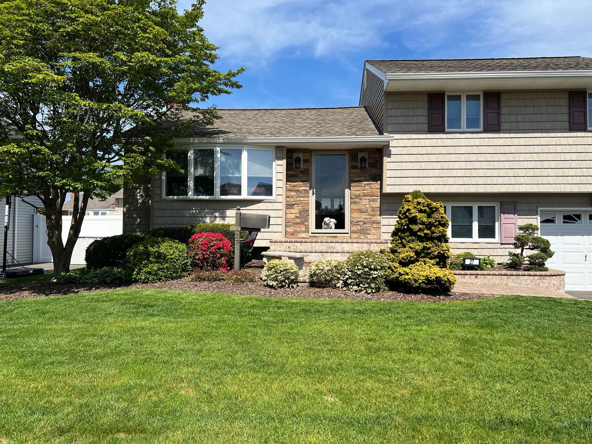 A split-level suburban house with beige siding, a stone entryway, and a front yard with trees and shrubs.