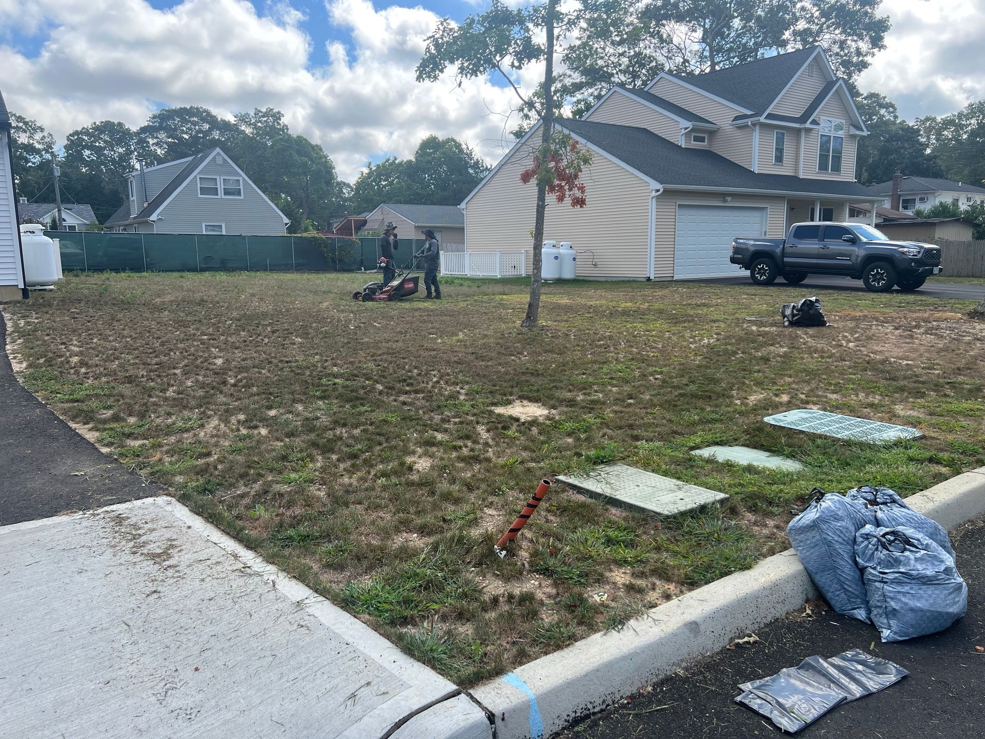 Two people work on a grassy residential lot near a sidewalk, with a house and a pickup truck in the background.
