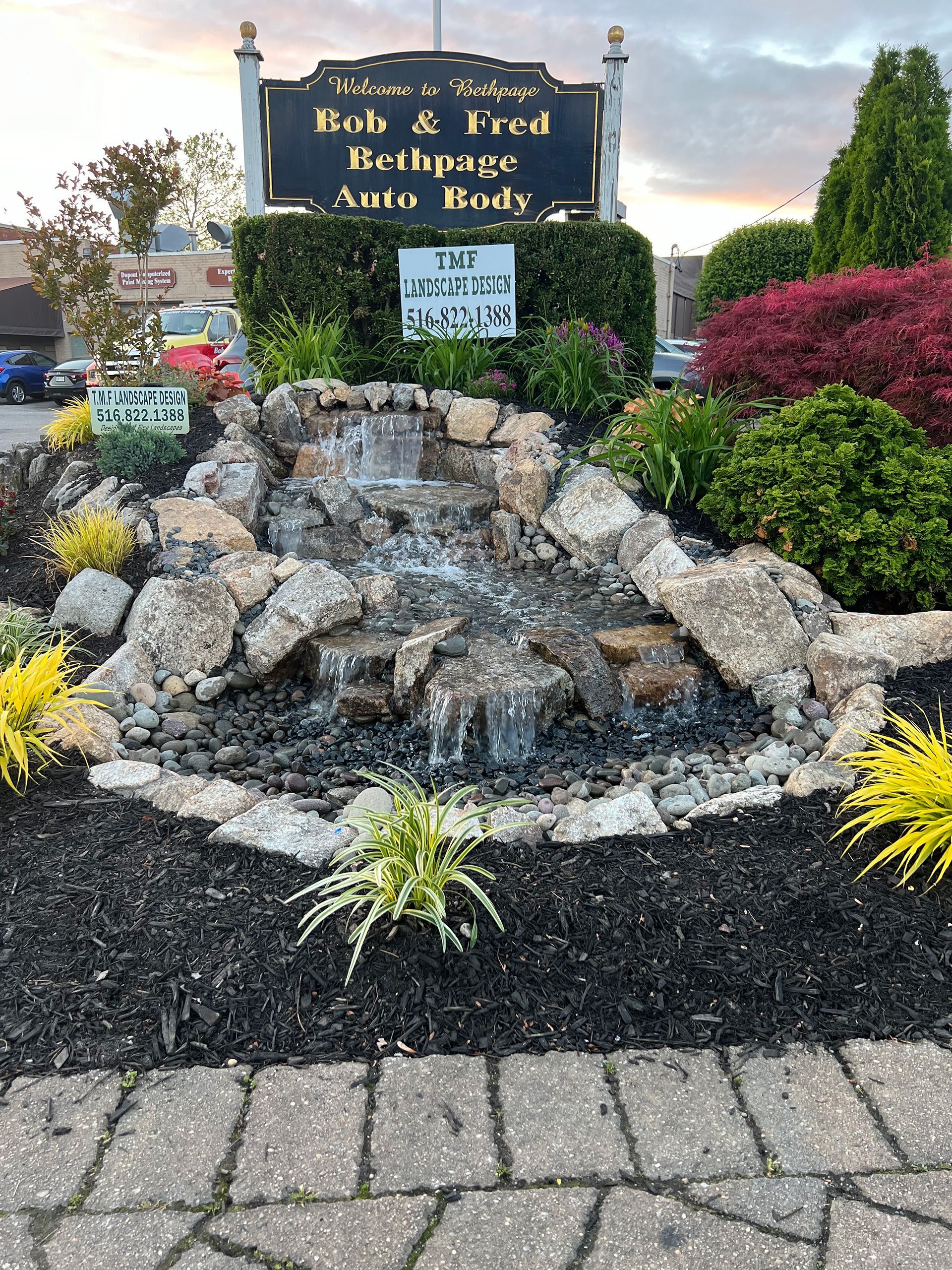 A sign for Bob & Fred Bethpage Auto Body sits above a tiered stone waterfall surrounded by landscaping and dark mulch.