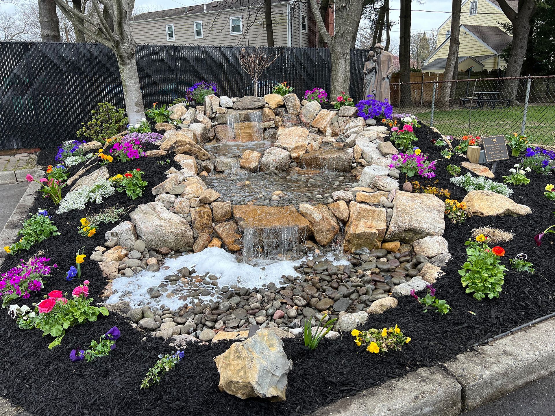 A tiered backyard water feature with cascading water, surrounded by tan rocks, black mulch, and colorful spring flowers.