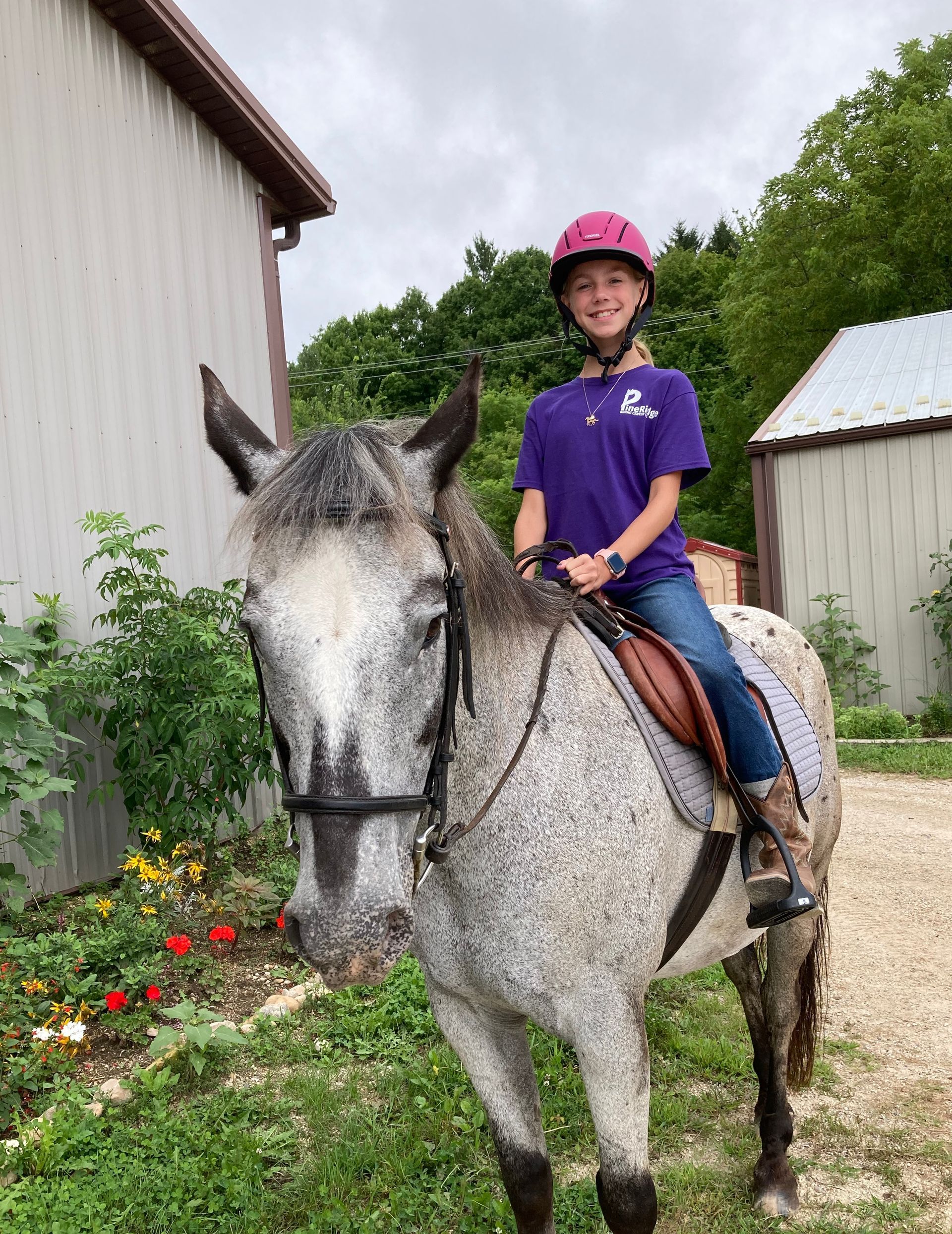 Girl in pink helmet smiles, riding speckled gray horse near barns.