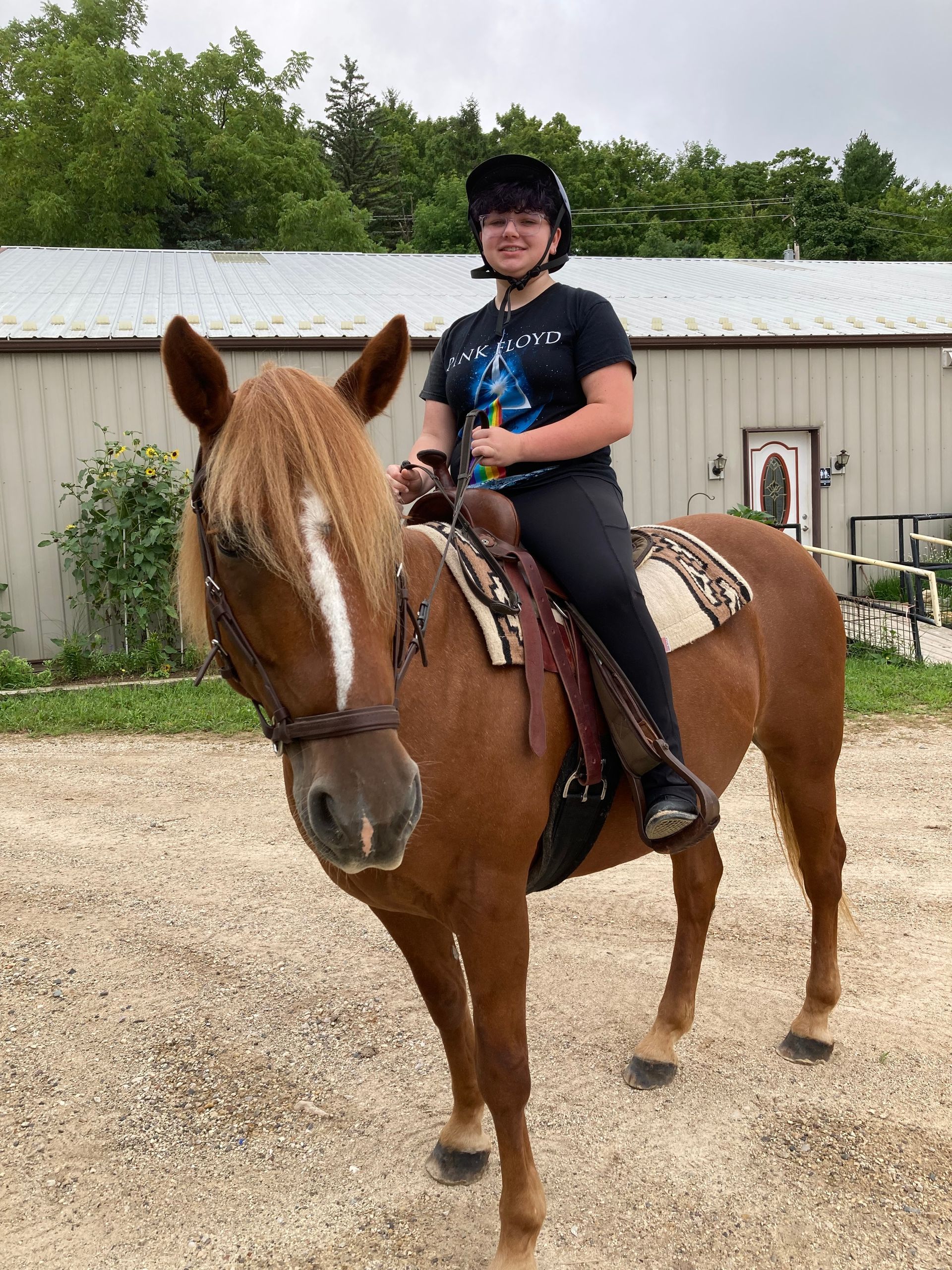 Person smiling on a tan horse with white blaze, wearing helmet and riding clothes, outdoors by a building.
