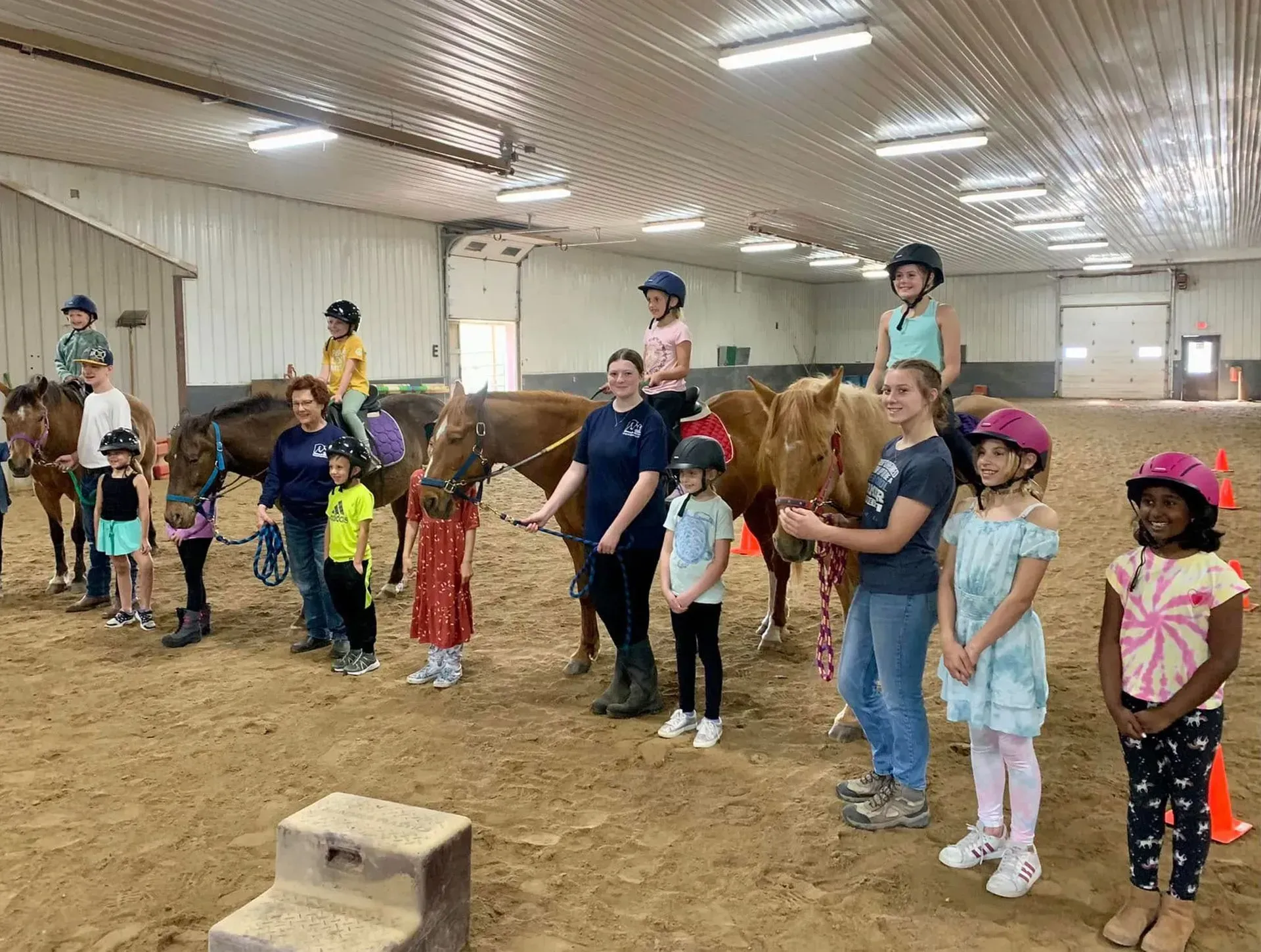 Kids and adults at an indoor horse riding lesson. People stand near horses with some riding them.