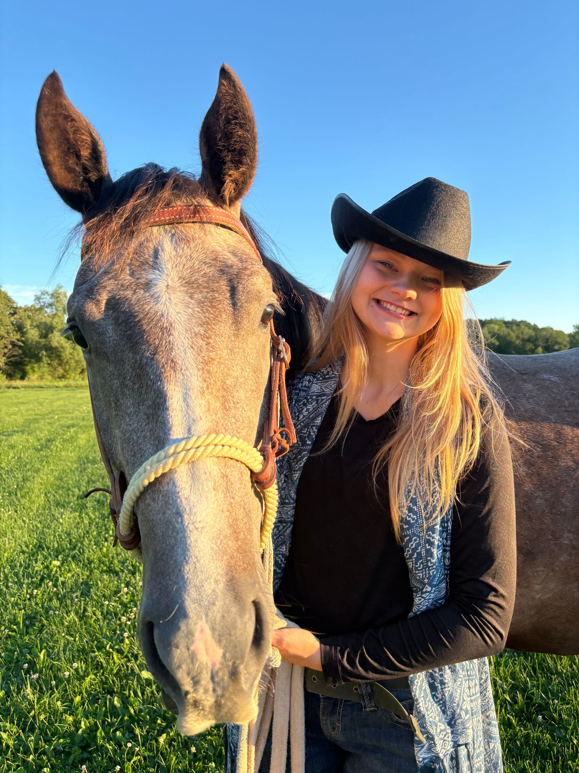 Woman in a cowboy hat smiles beside a gray horse in a sunny field.