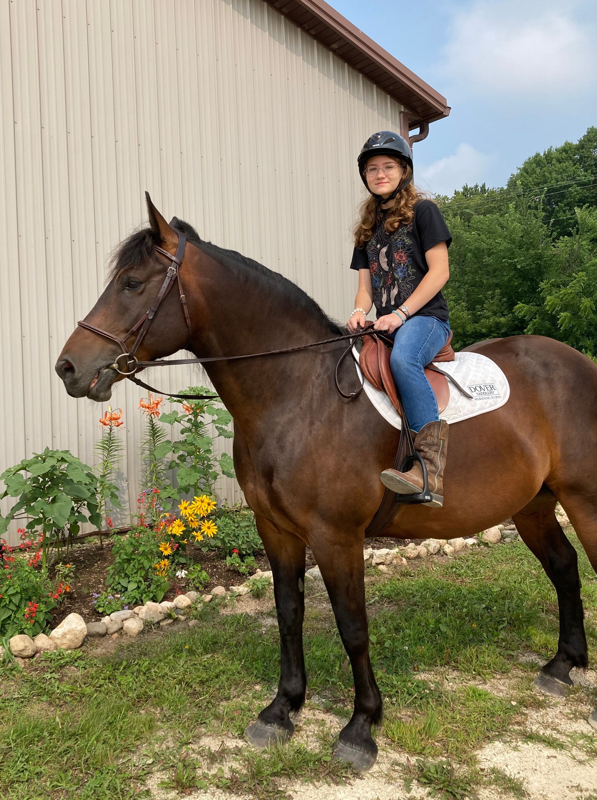 Girl in helmet riding a brown horse in front of a tan building and greenery.