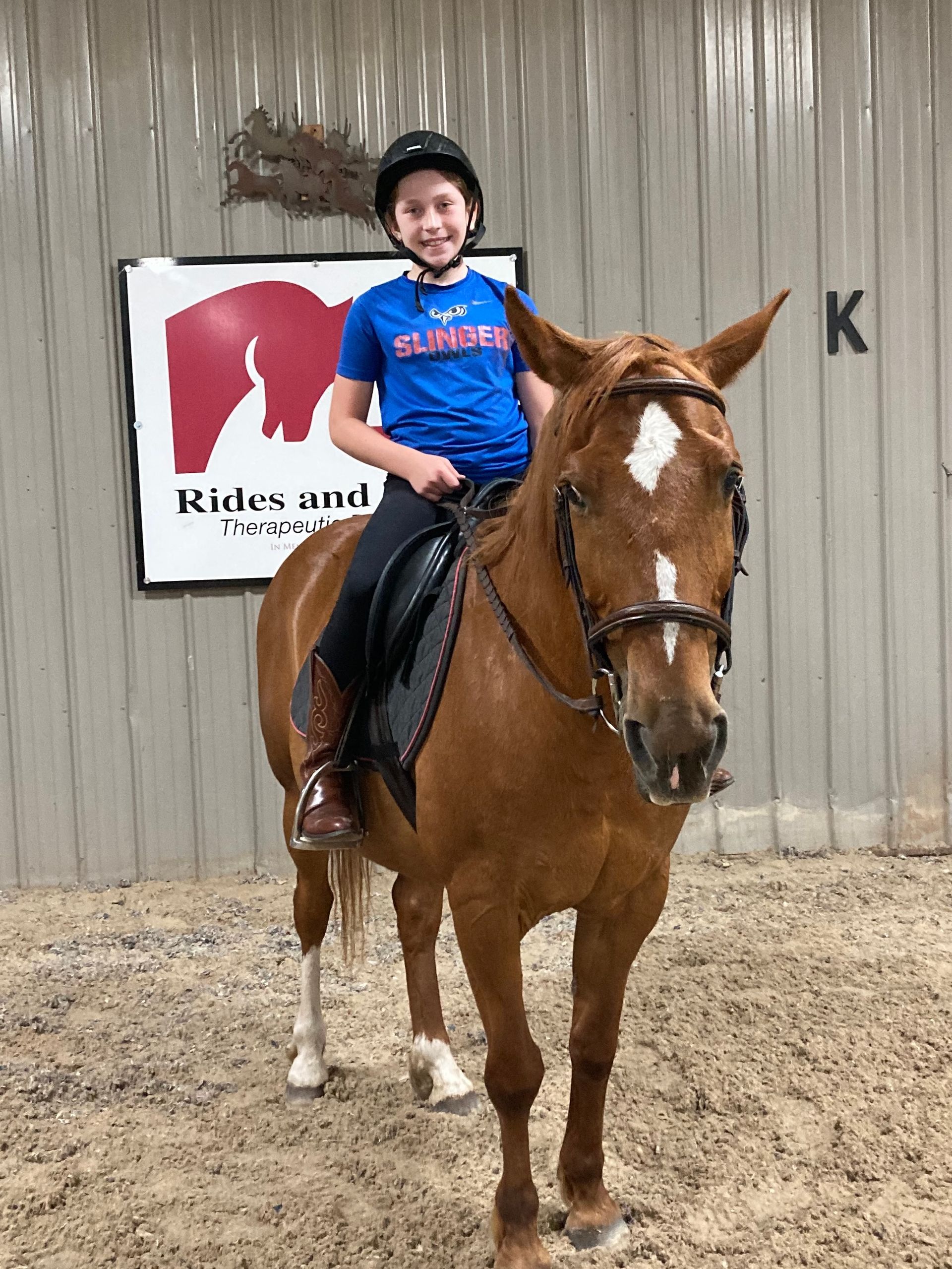 Young person on a chestnut horse in an indoor arena, smiling, wearing a helmet. 