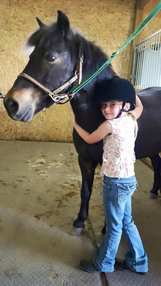 Girl in riding helmet hugging a dark horse in a stable.