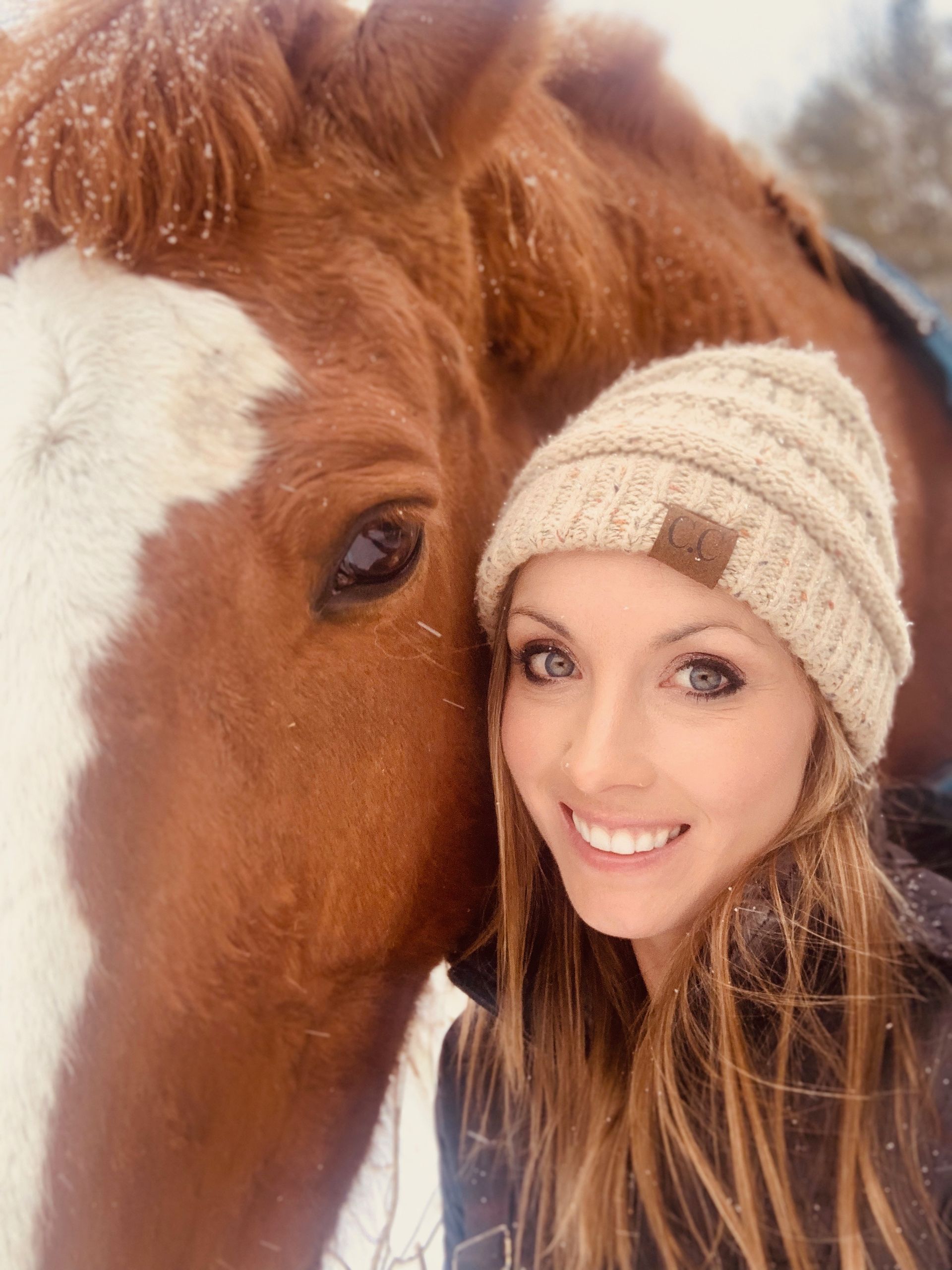 Woman in beanie smiles, posed beside a brown horse with a white face marking, in snowy setting.