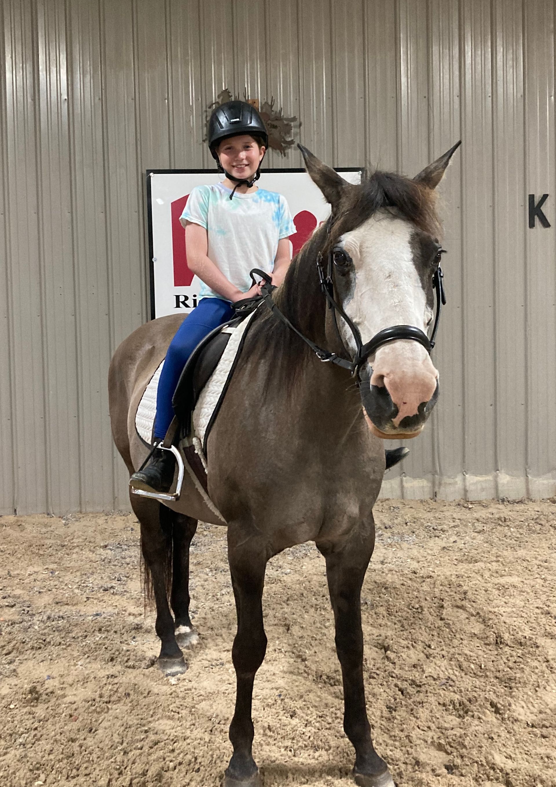 Girl in riding gear on a gray horse in an indoor arena. Horse has a white face, rider smiles.
