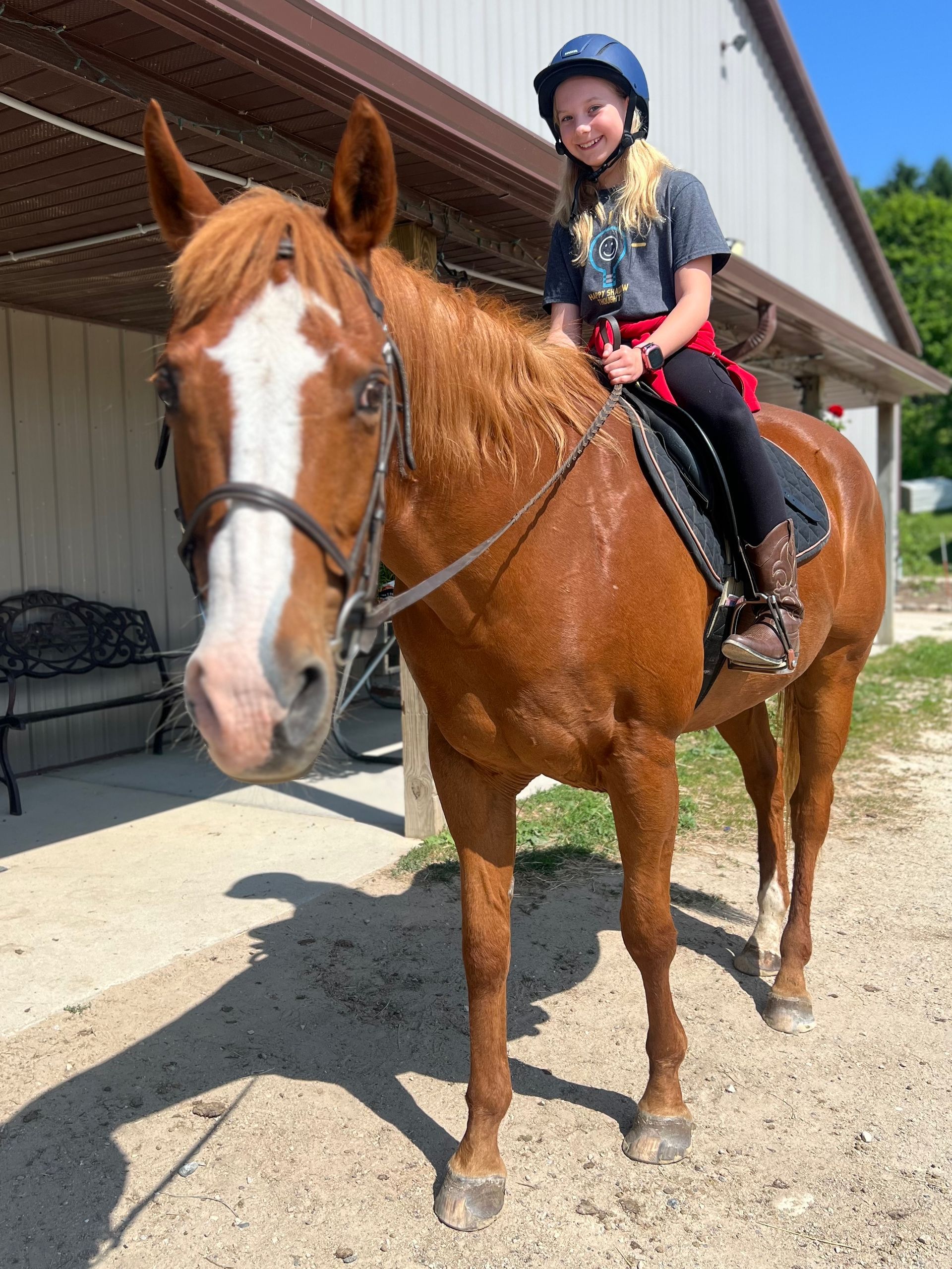 Young person with a blue helmet smiles atop a chestnut horse with a white face marking, near a building.