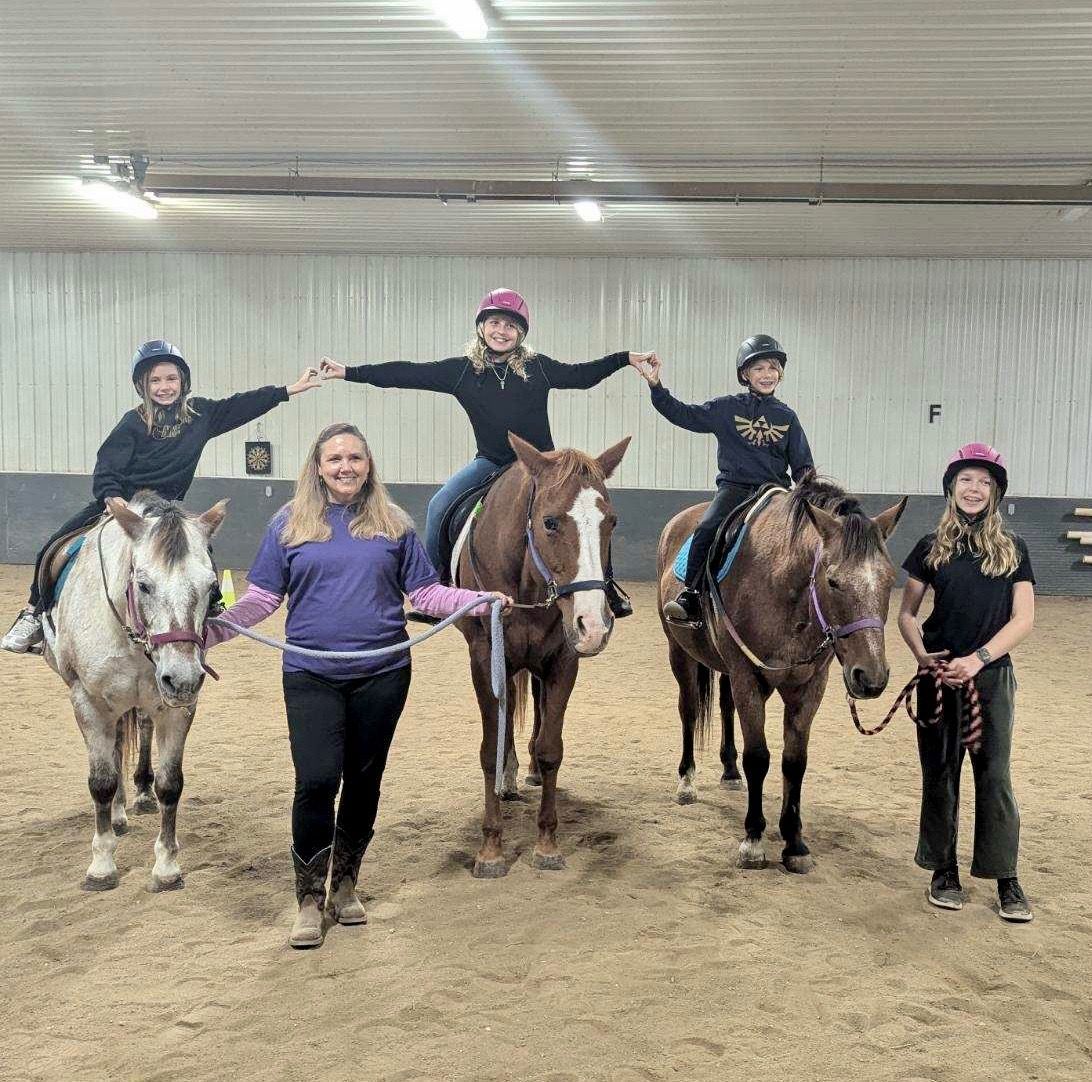 Group of kids and an adult posing with horses in an indoor riding arena.