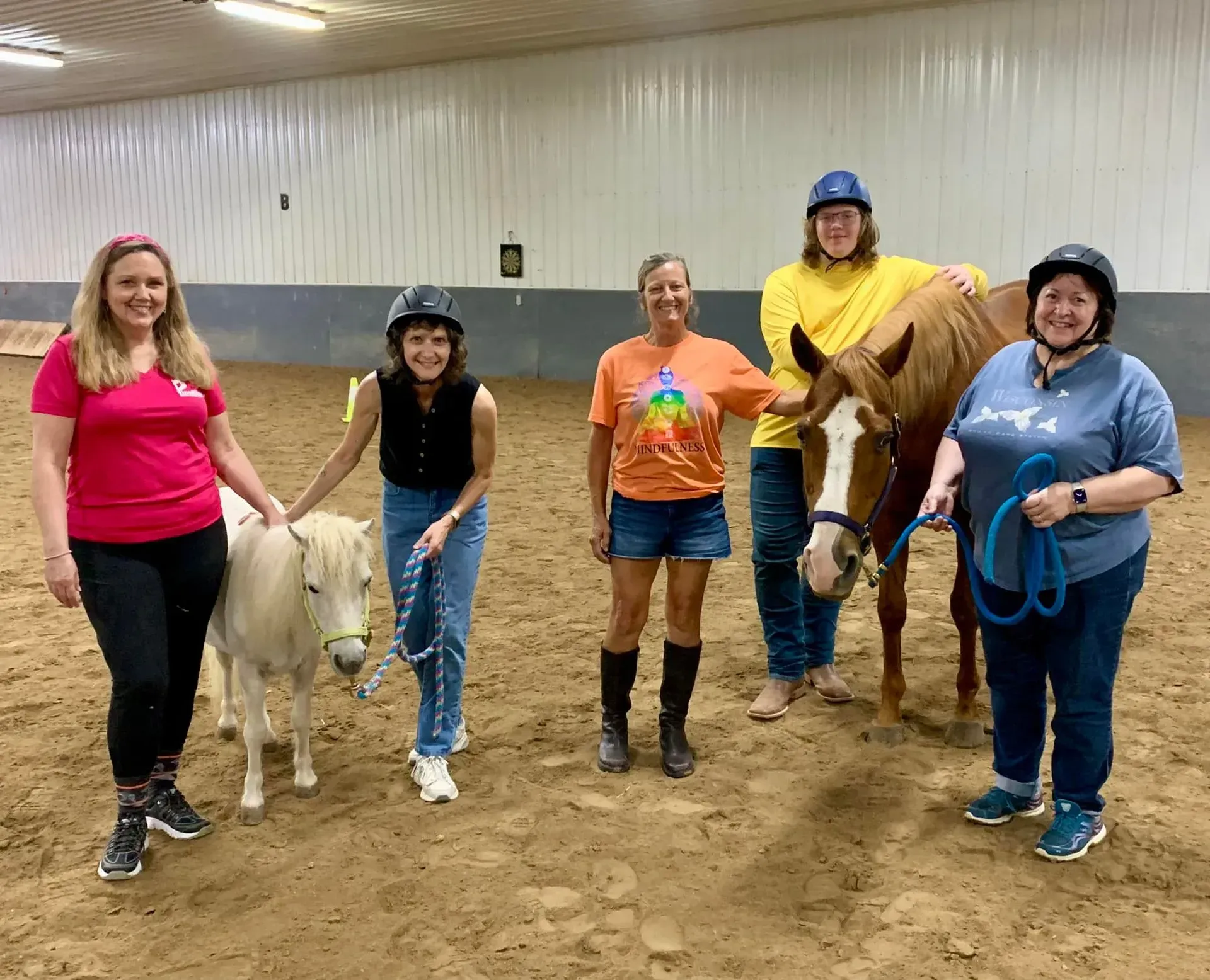 Five people with two horses in an indoor arena. People are smiling, one pats a miniature horse.