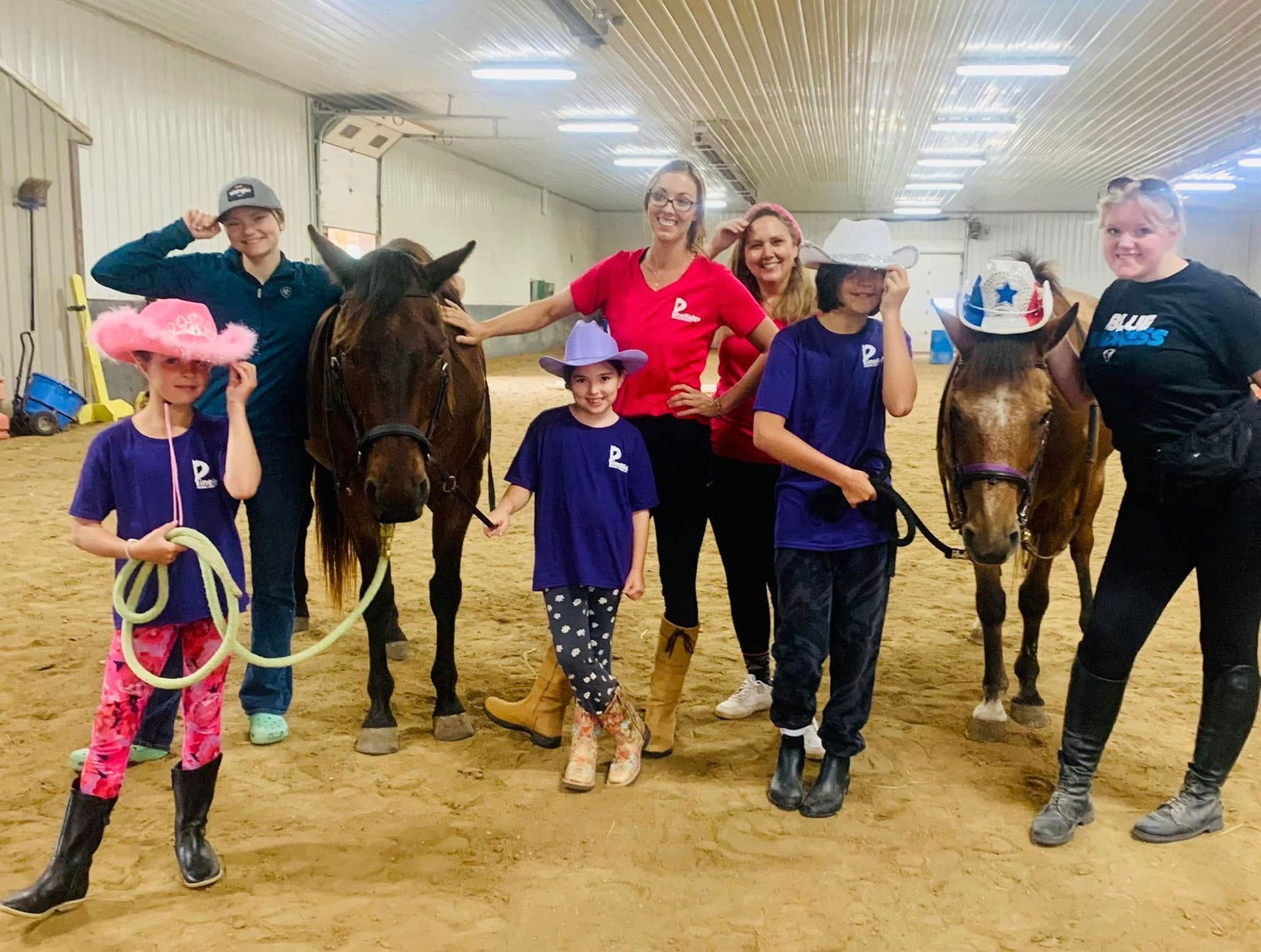 People and horses in an indoor arena. Some wear cowboy hats, smiling. One holds a rope.
