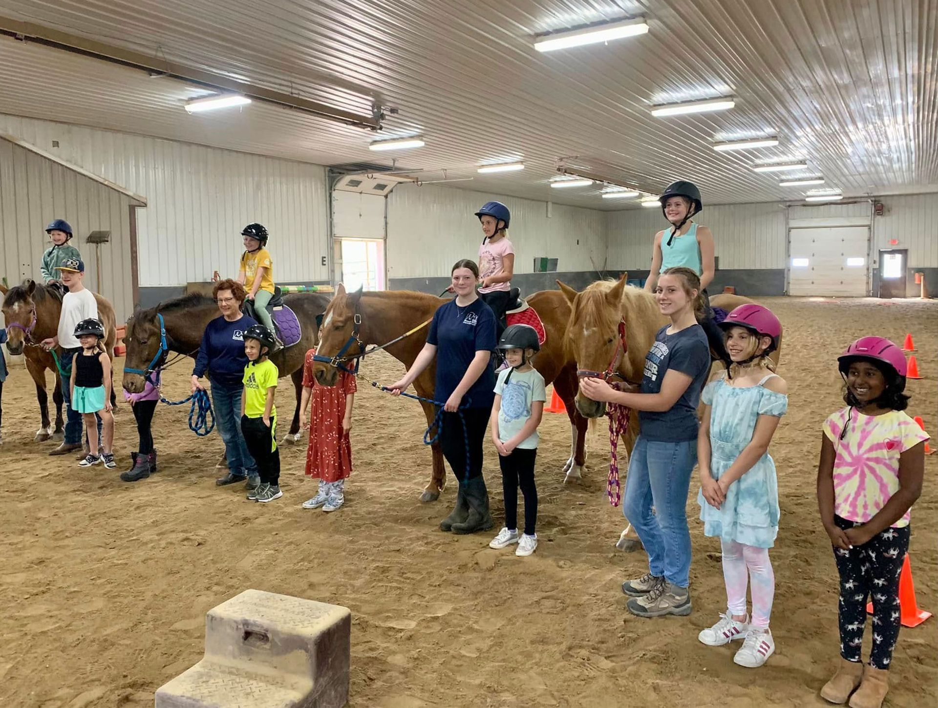Children and adults in an indoor riding arena with horses, some riding, others standing.