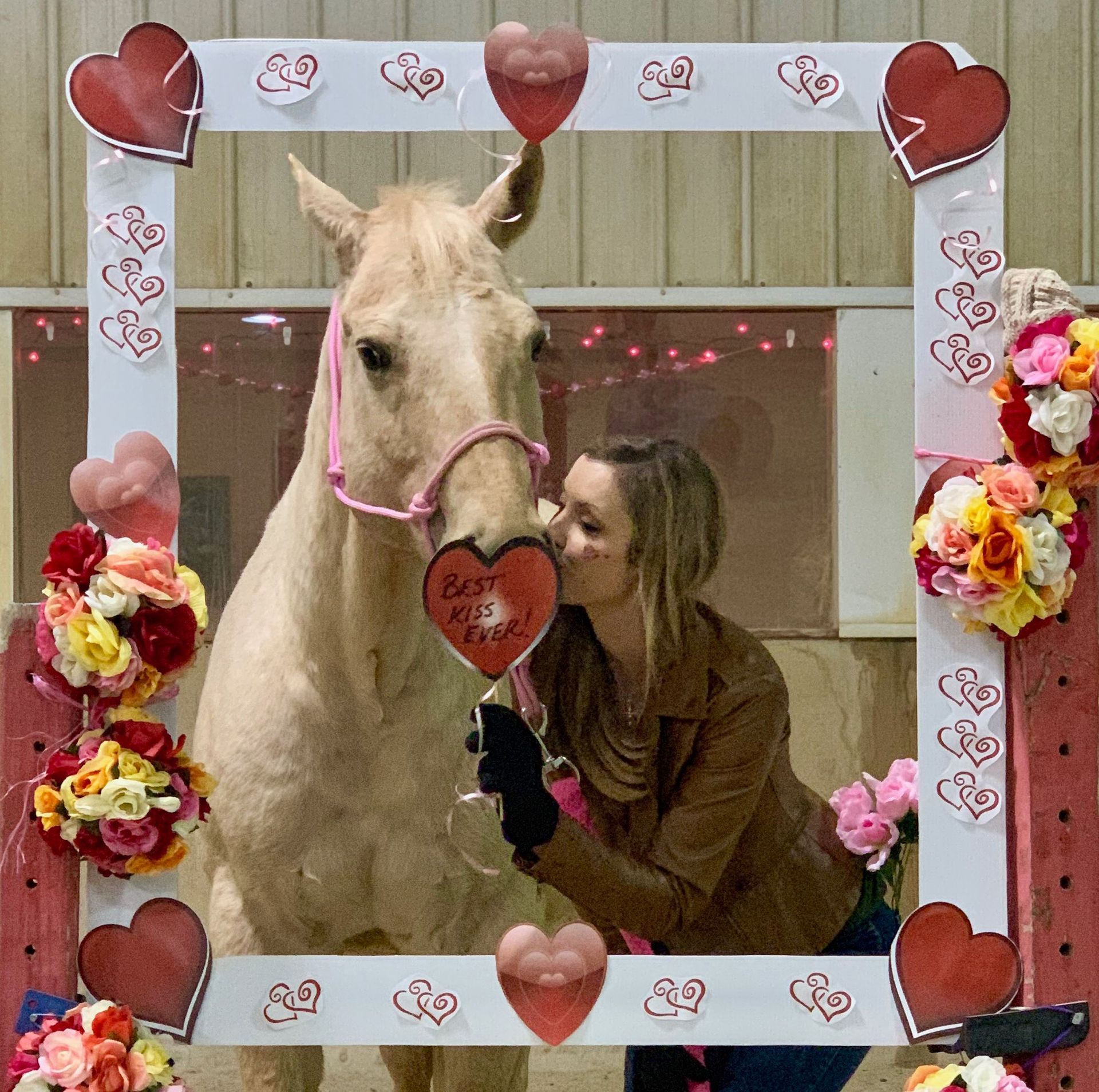 Woman kissing a pale horse through a Valentine's Day photo frame. Balloons and bouquets decorate the frame.
