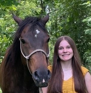 Woman with long brown hair smiles beside a brown horse, both looking at the camera outdoors.