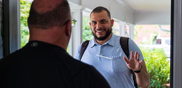 Man with backpack smiles and waves at a doorway, facing another person indoors.