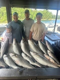 Four people stand behind a table piled with large striped fish at a dockside fish-cleaning area.