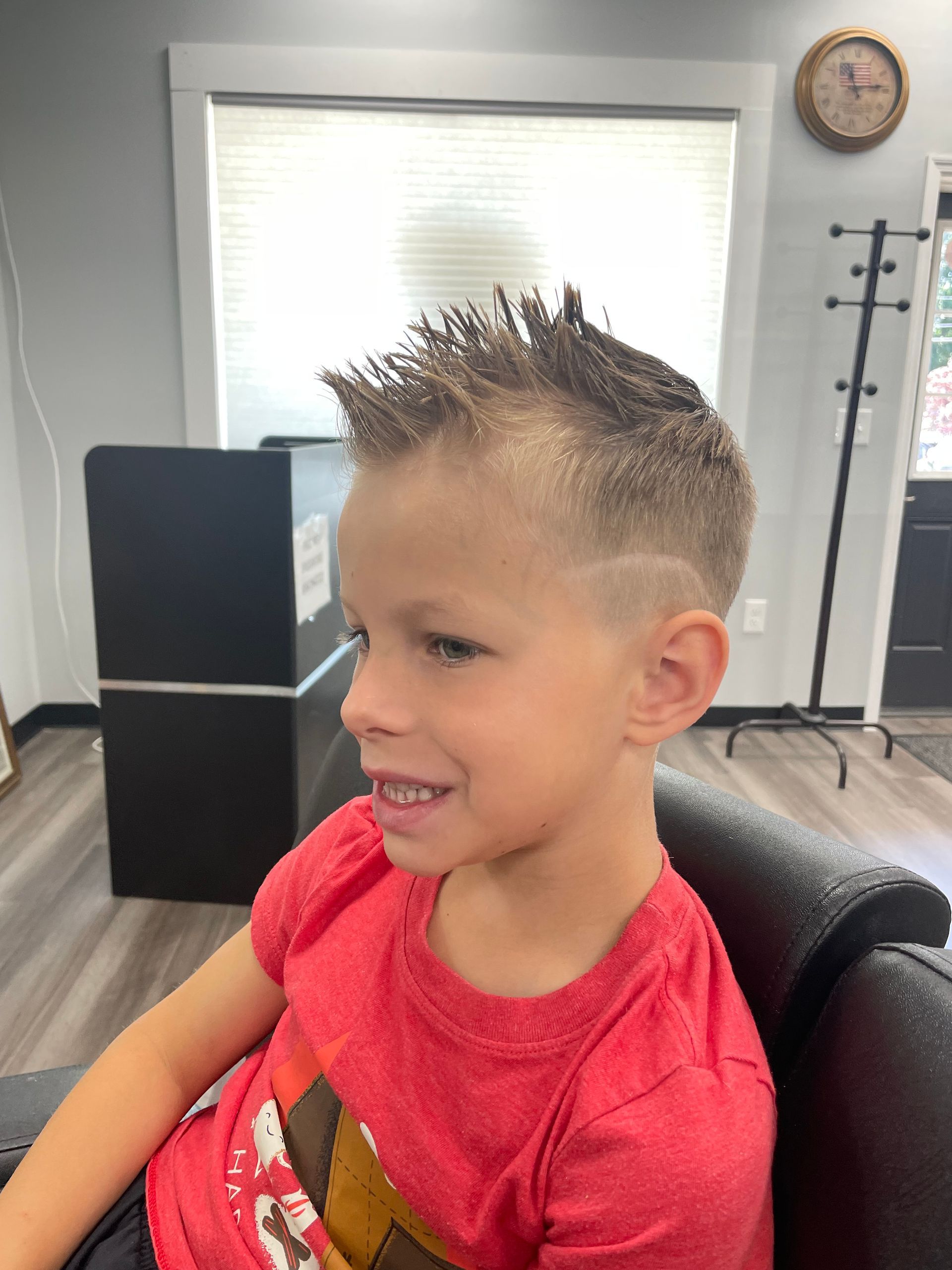 A young boy in a red shirt is sitting in a chair in a barber shop.