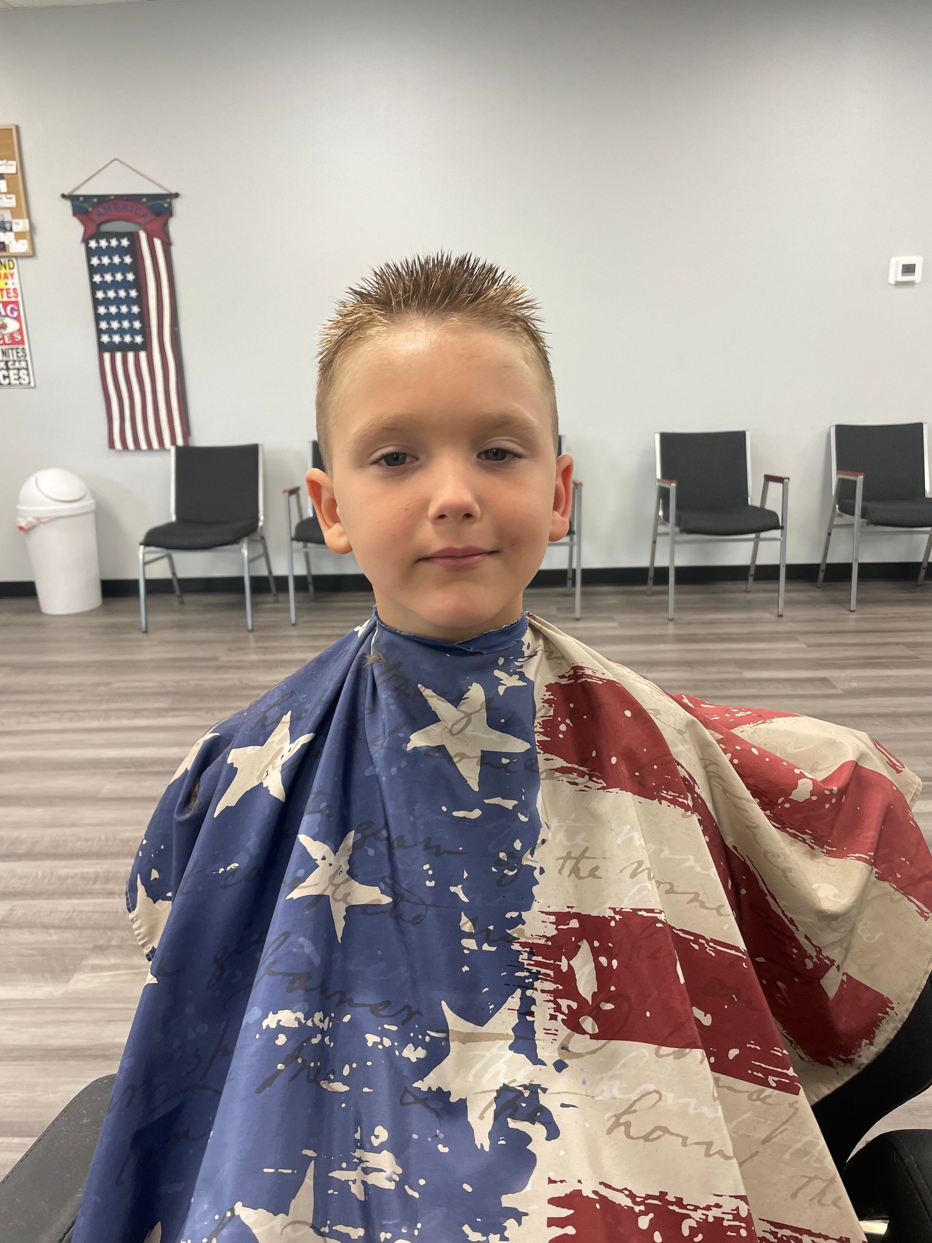 A young boy is sitting in a chair wearing an american flag barber cape.