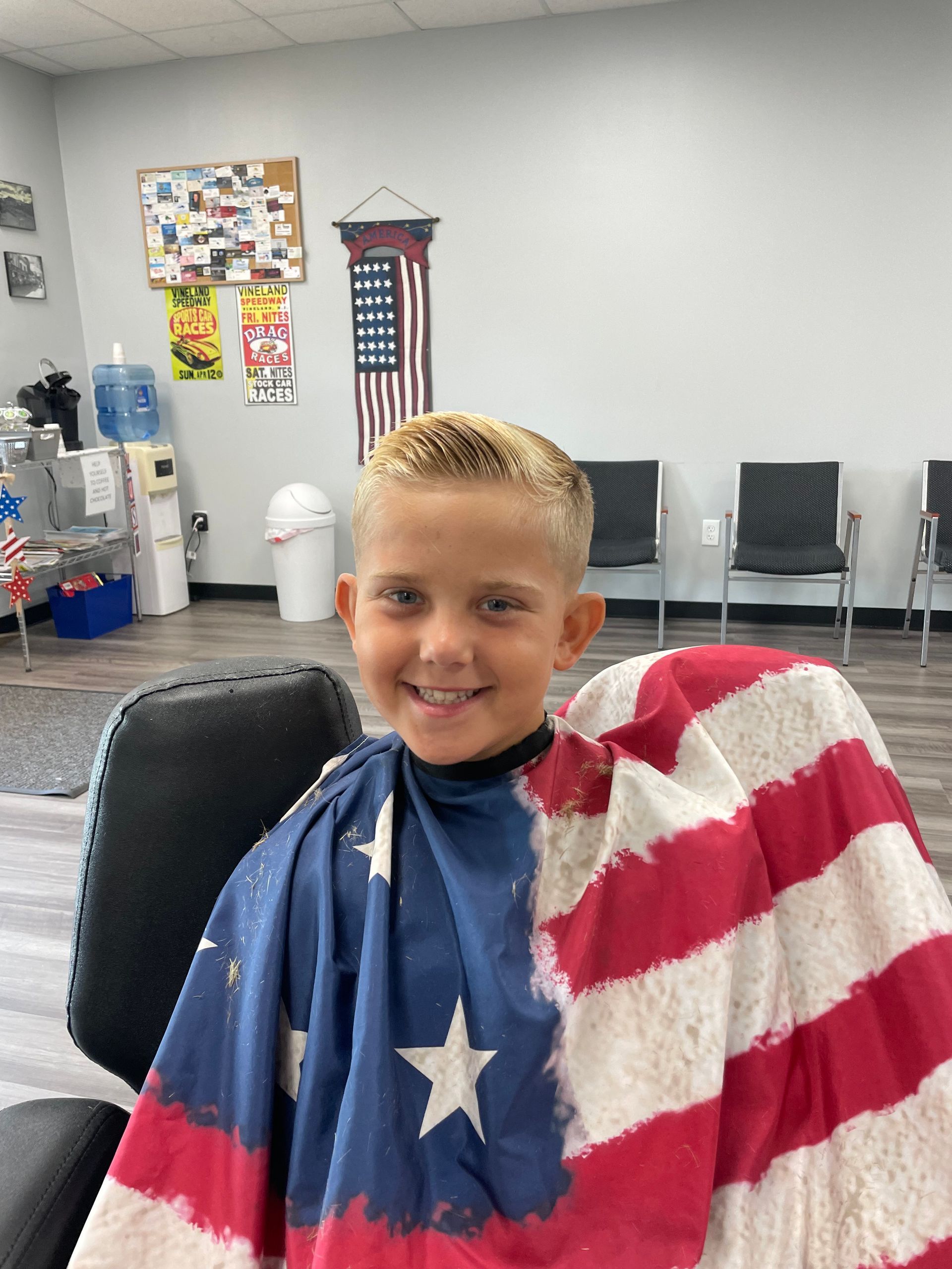 A young boy is sitting in a chair wearing an american flag barber cape.
