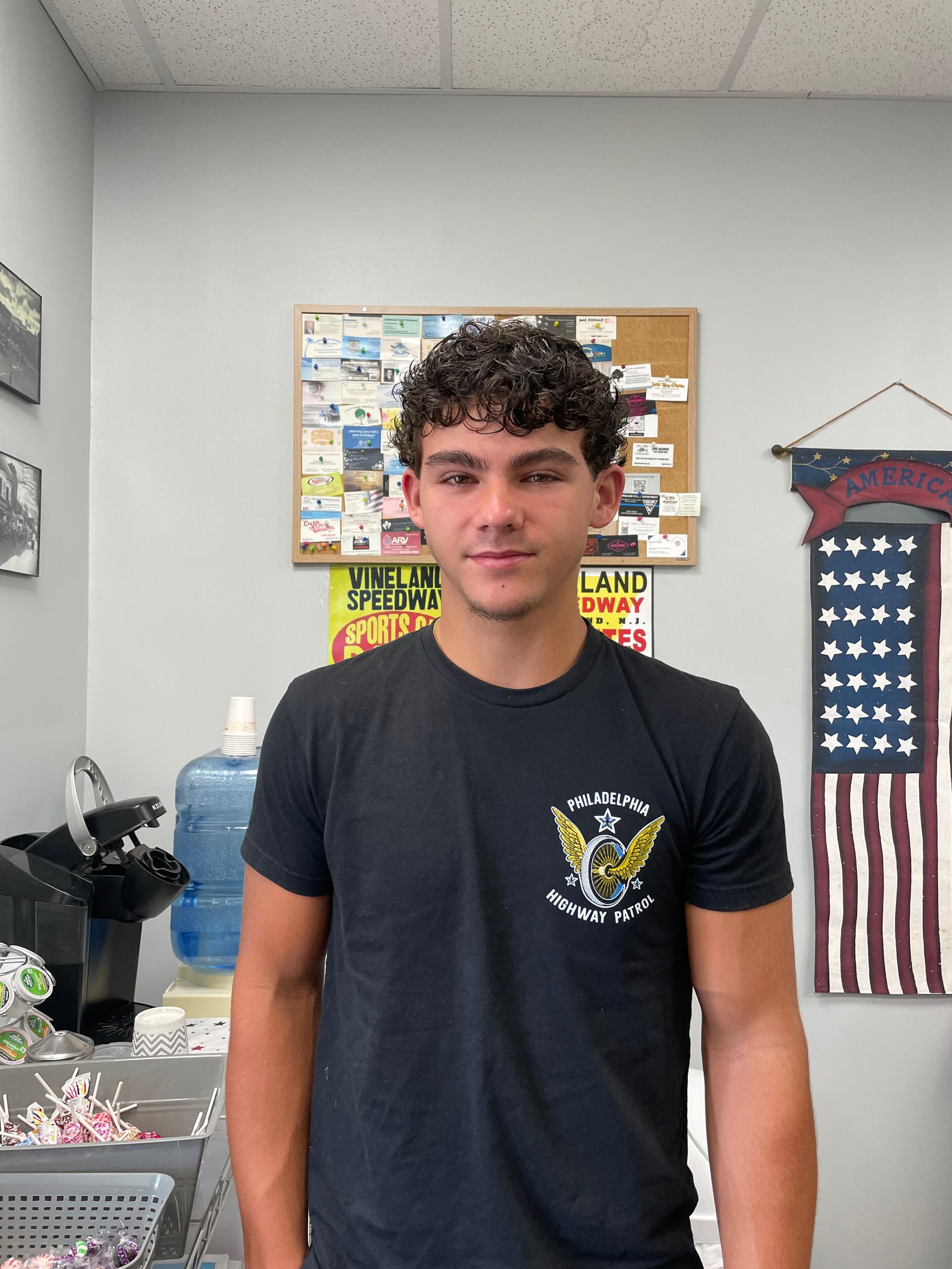 A young man in a black shirt stands in front of an american flag