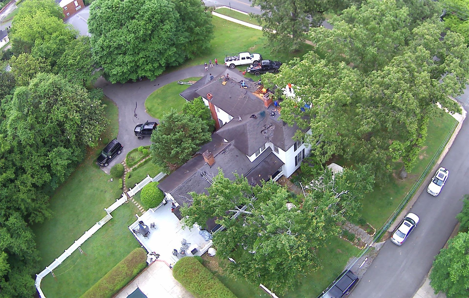 An aerial view of a house with a dark shingled roof, surrounding trees, a driveway with parked cars, and a paved street.