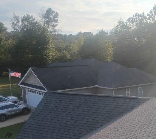 An elevated view of a house roof, a garage with an American flag, a parked vehicle, and a tree-lined background.