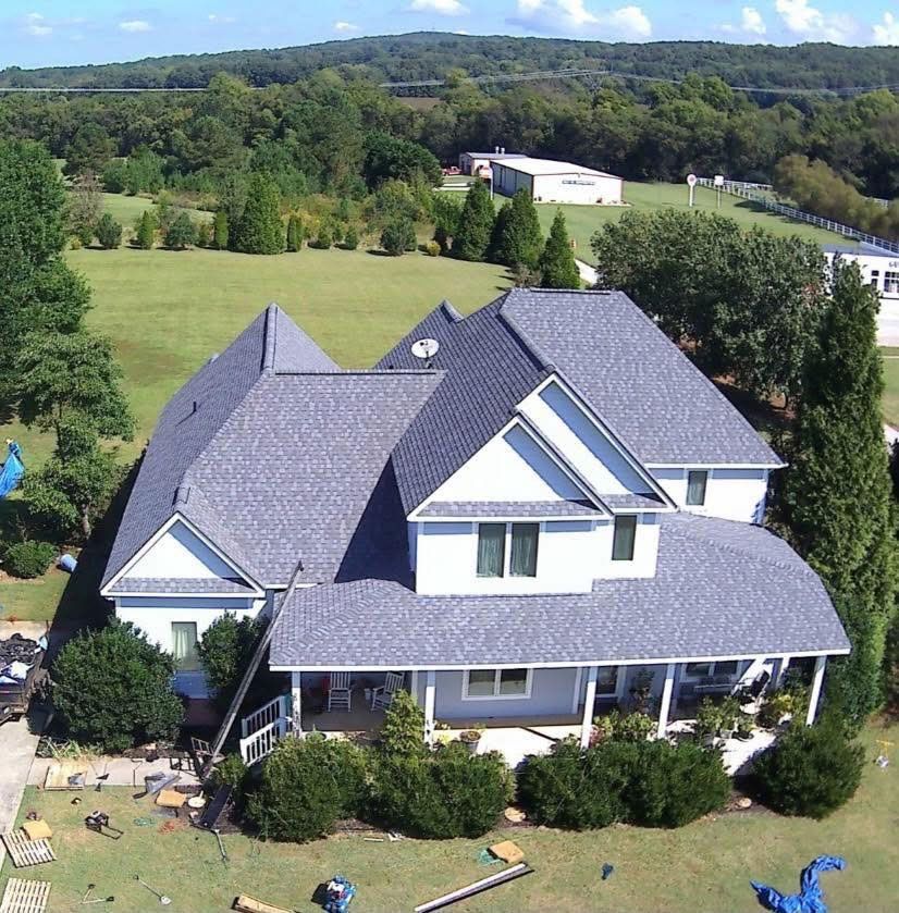 Aerial view of a white two-story house with a grey shingled roof, surrounded by green lawns and trees on a sunny day.