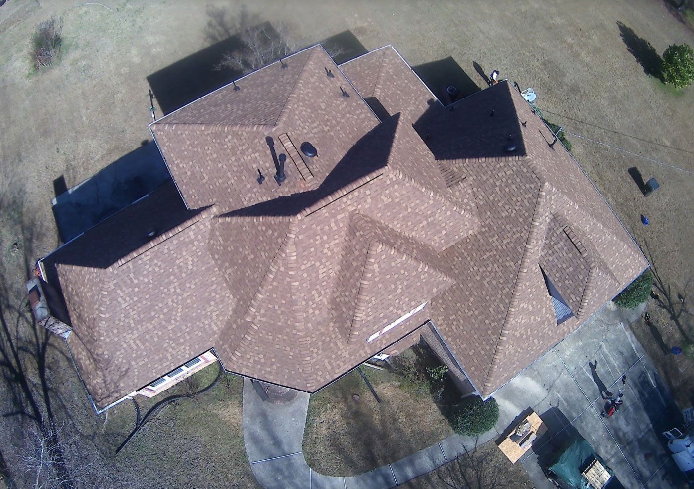 An overhead drone view of a large residential house with a complex brown shingled roof, set in a yard.