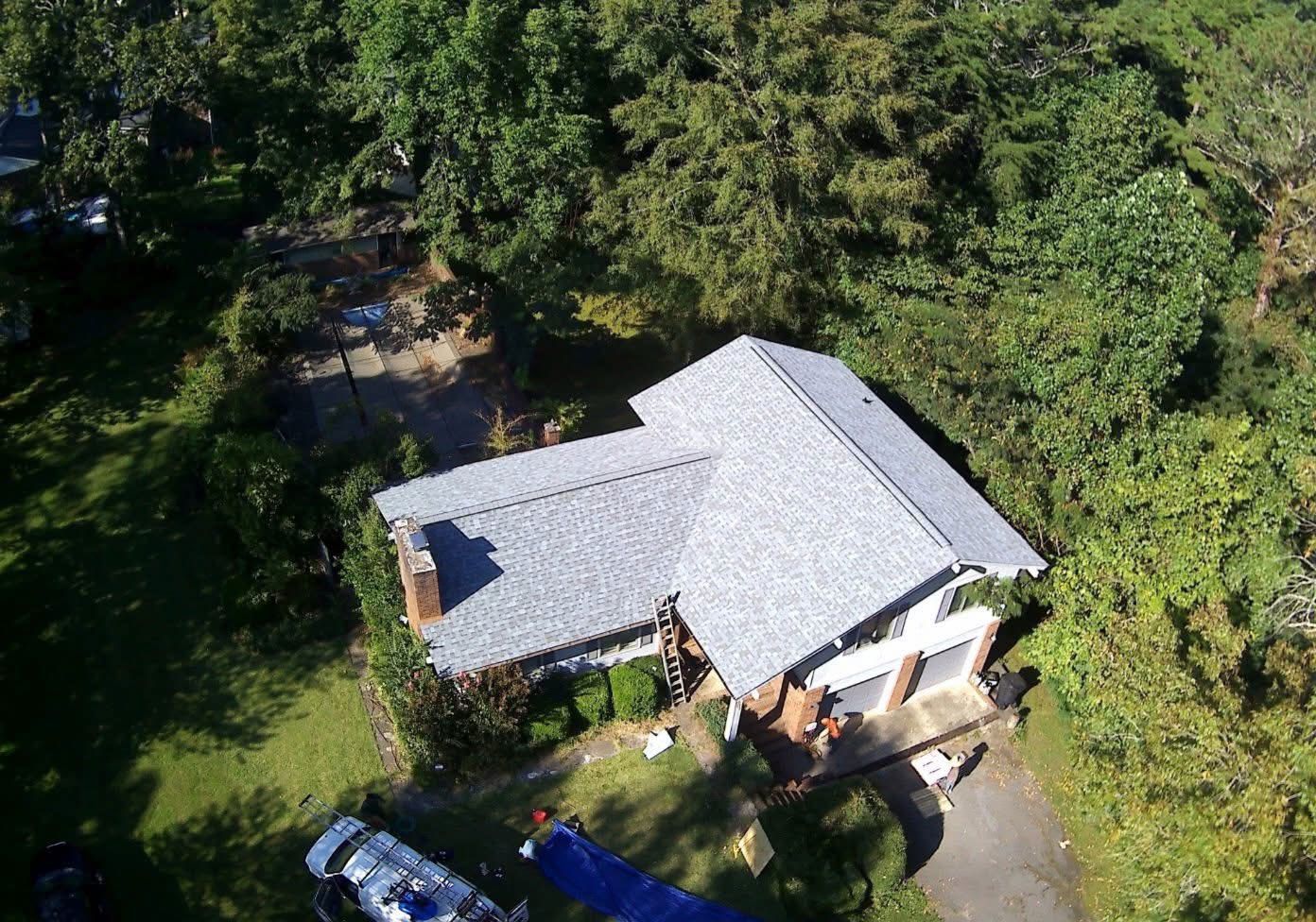Aerial view of a house with a gray shingled roof, surrounded by trees and a lawn with a vehicle and a blue tarp.