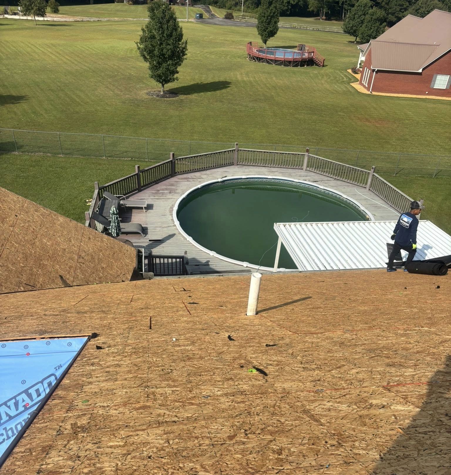 A worker on a roof under construction looks down at a pool with a green cover, a wooden deck, and a yard.