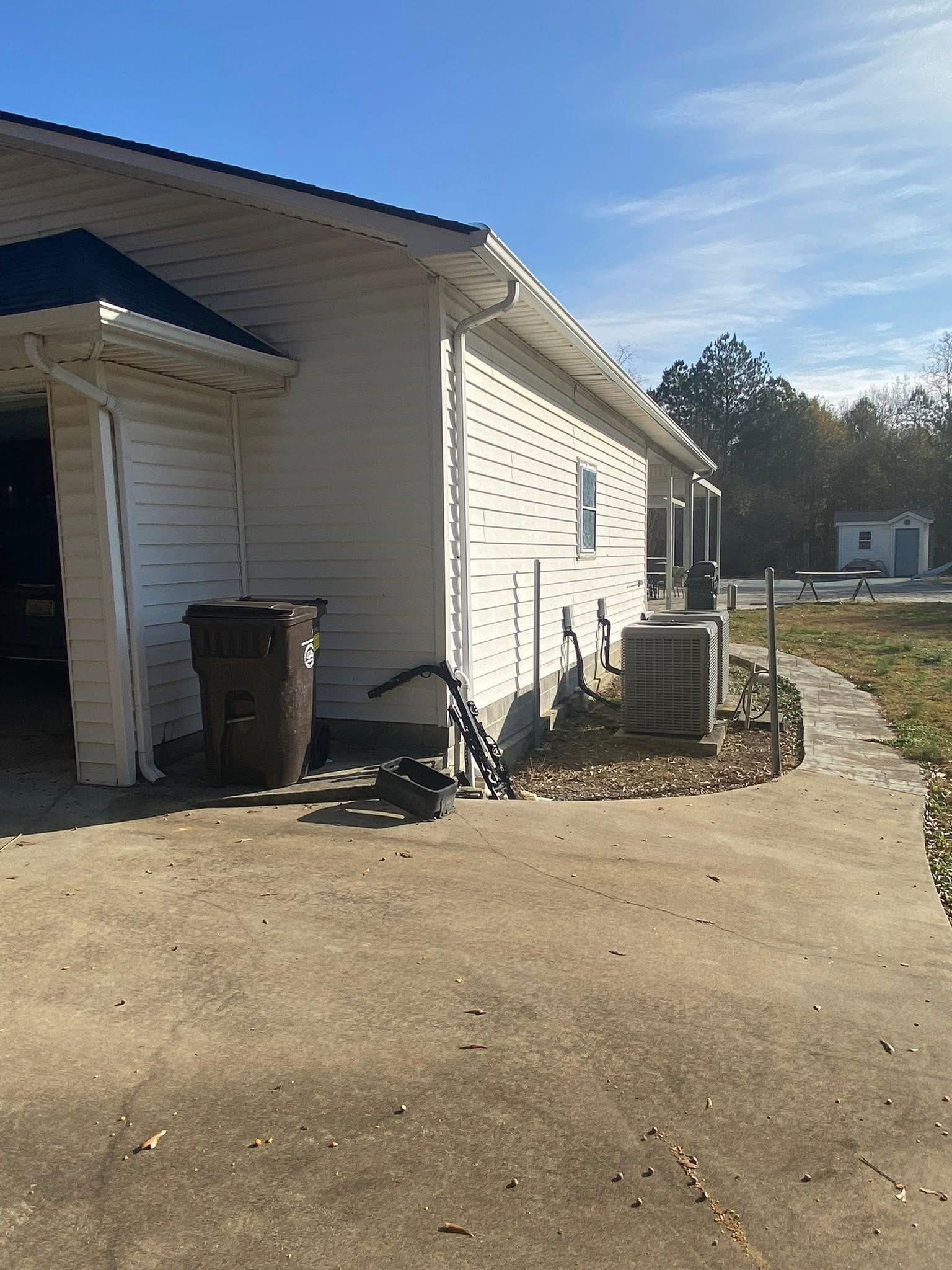 A white house exterior featuring a trash bin, an HVAC unit, and a paved walkway under a clear blue sky.