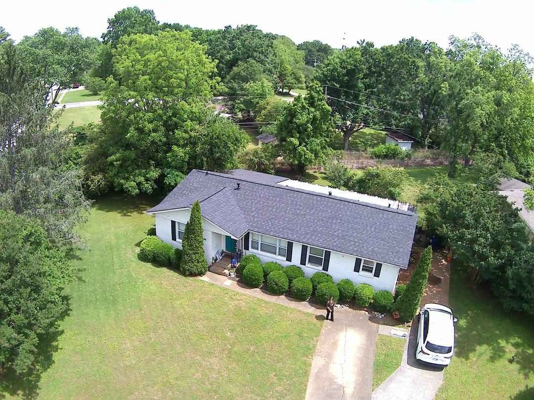 Aerial view of a white single-story house with a dark roof, surrounded by trees and a green lawn with a parked white car.