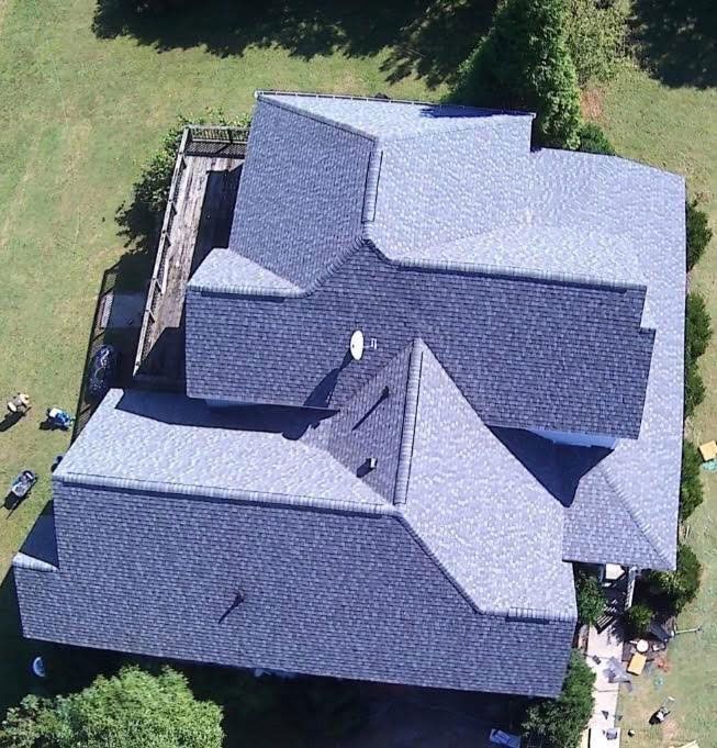 Aerial view of a complex, multi-gabled gray shingled roof on a suburban house surrounded by green lawn and trees.