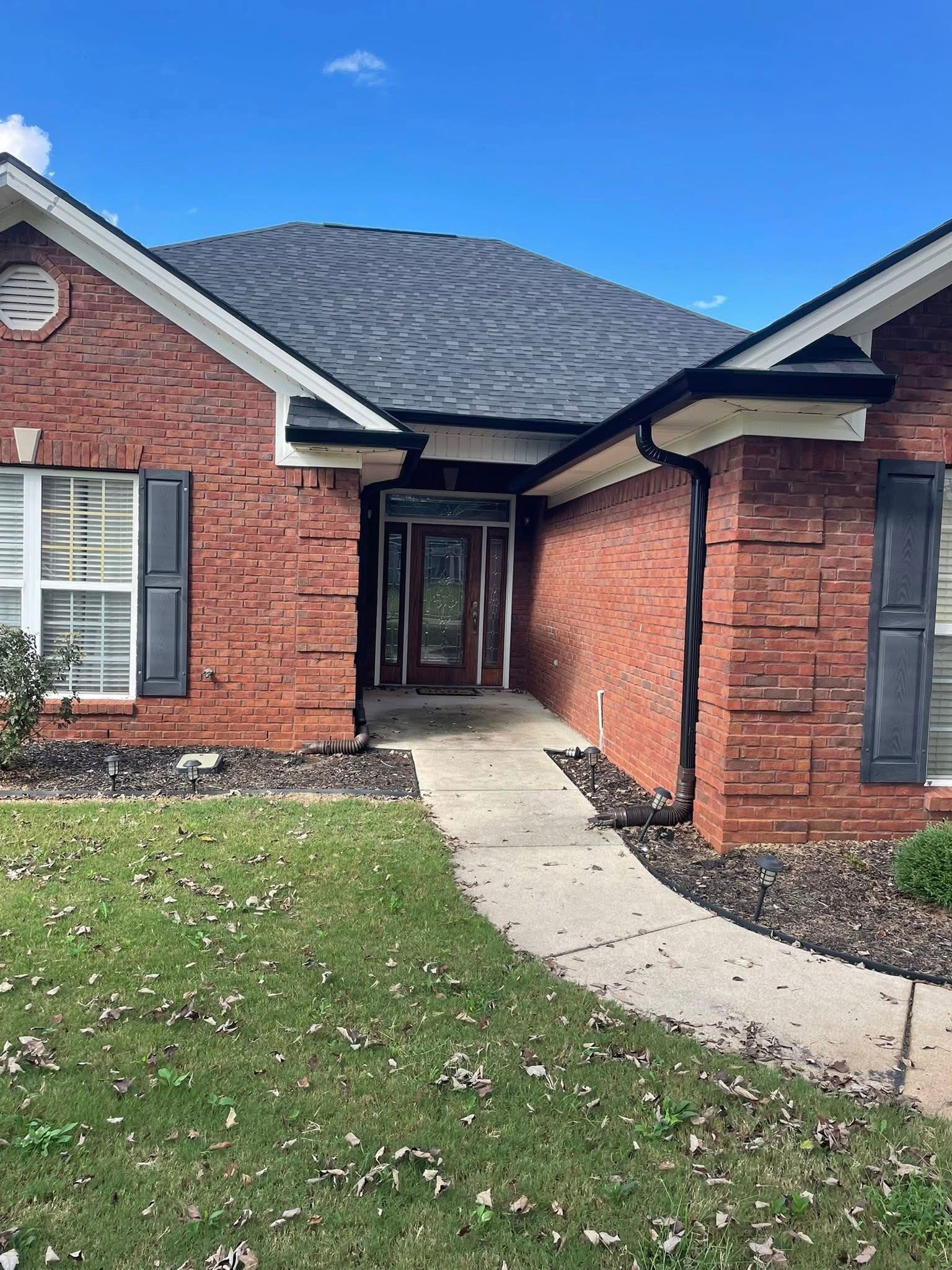 A red brick single-story house with a grey roof, dark shutters, and a concrete walkway leading to the front entryway.