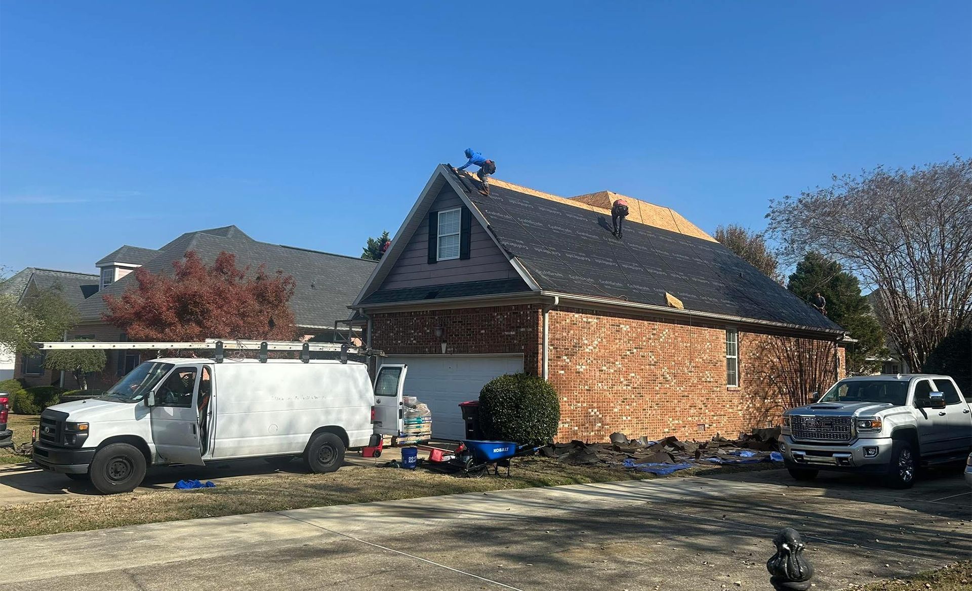 Workers repair the roof of a brick house on a sunny day, with a white van and a silver pickup parked nearby.