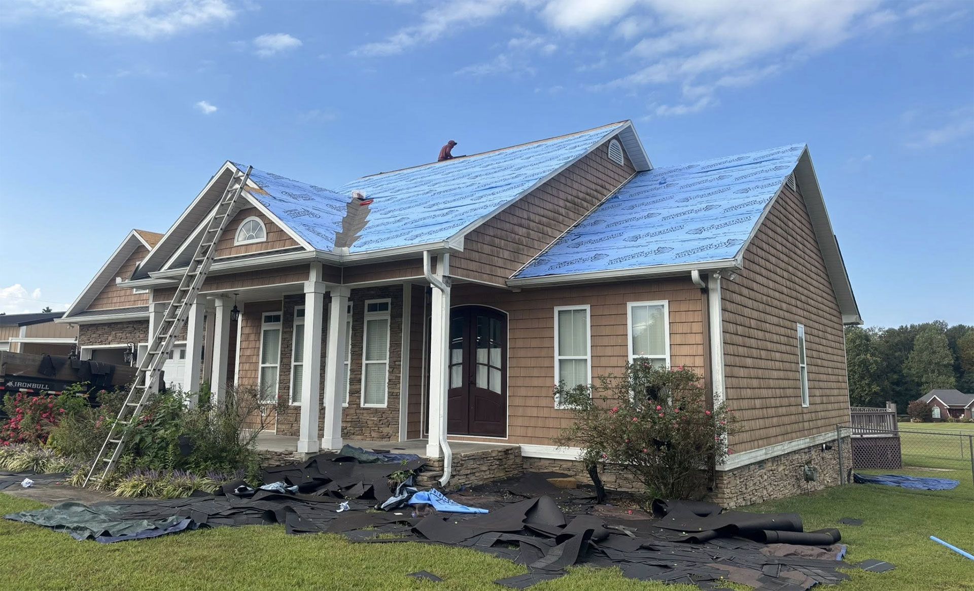 A tan brick house with columns and a bright blue roof underlayment being installed, with roofing debris on the lawn.