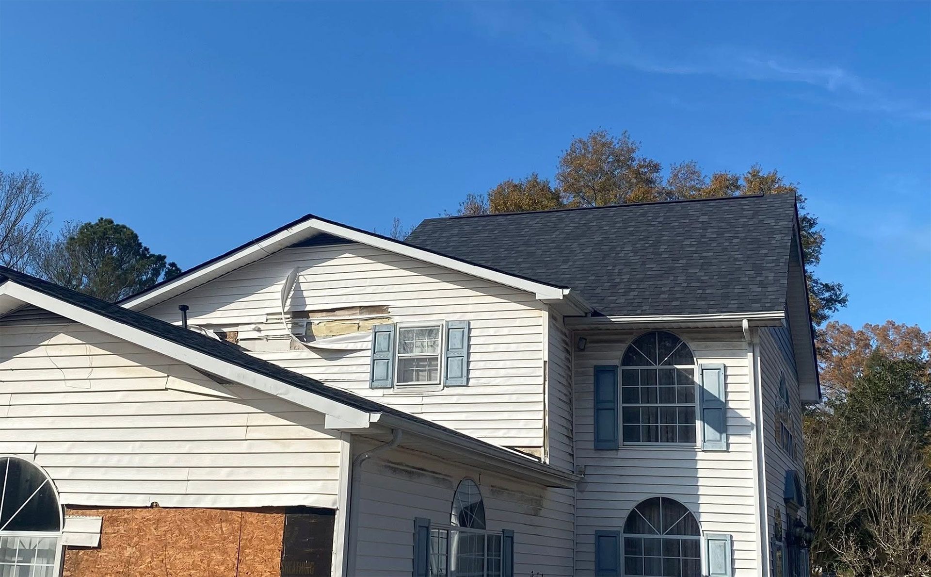 A house exterior with damaged, peeling white siding and a section of exposed wood near an upper-story window.