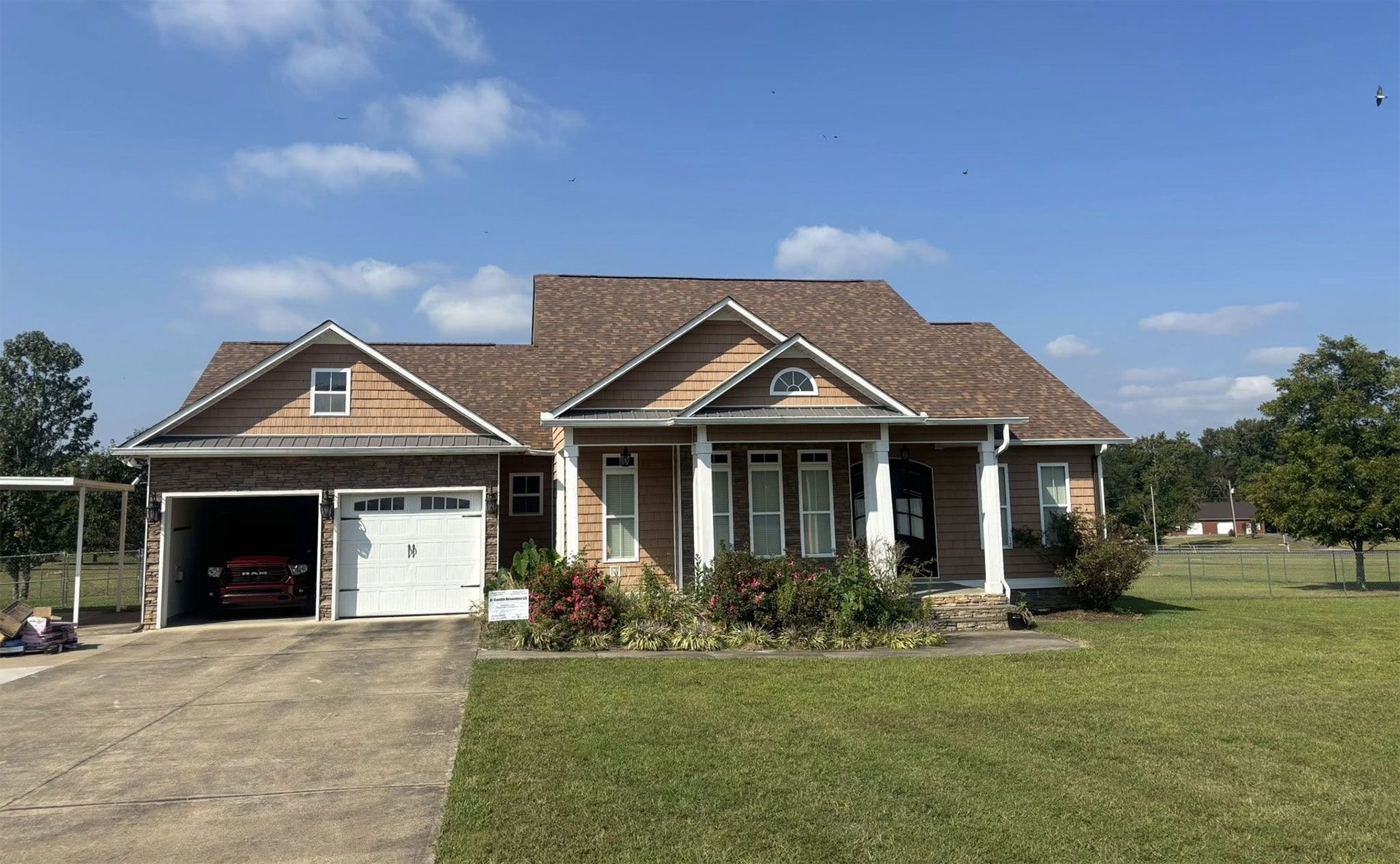 A single-story brick house with a front porch, columns, and a two-car garage, surrounded by a lawn under a blue sky.