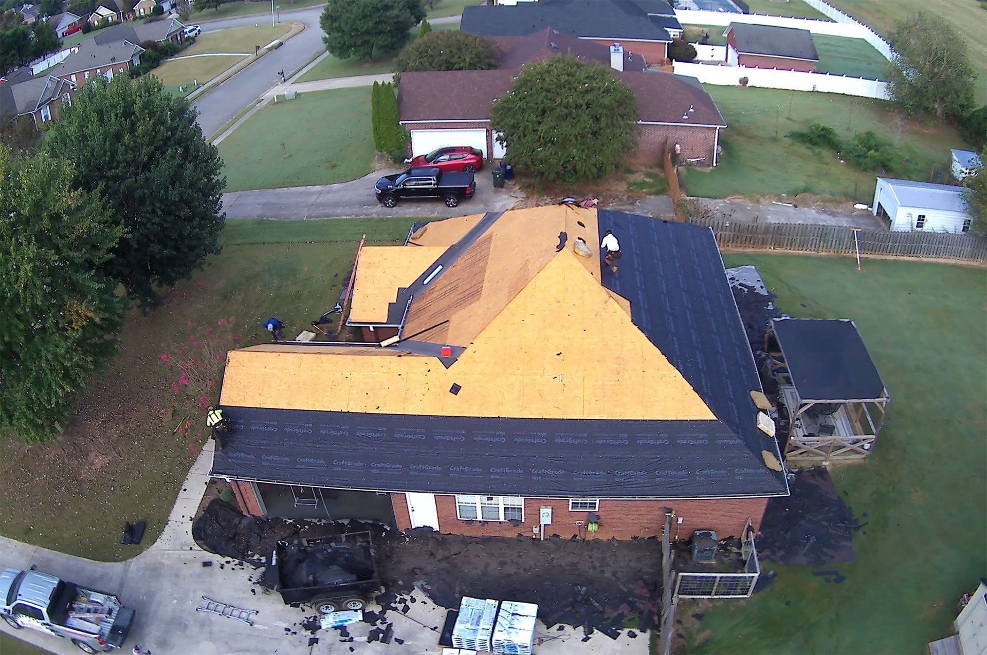 Aerial view of workers replacing shingles on a house roof, showing both exposed wood sheathing and completed sections.