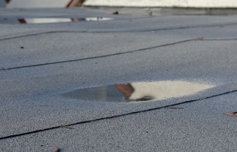 Puddles of water on a flat, gray roof; the sky and building reflected.