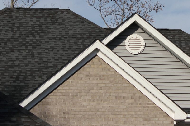 Close-up of a house roof with dark gray shingles, white trim, and a gray brick facade with an oval vent.